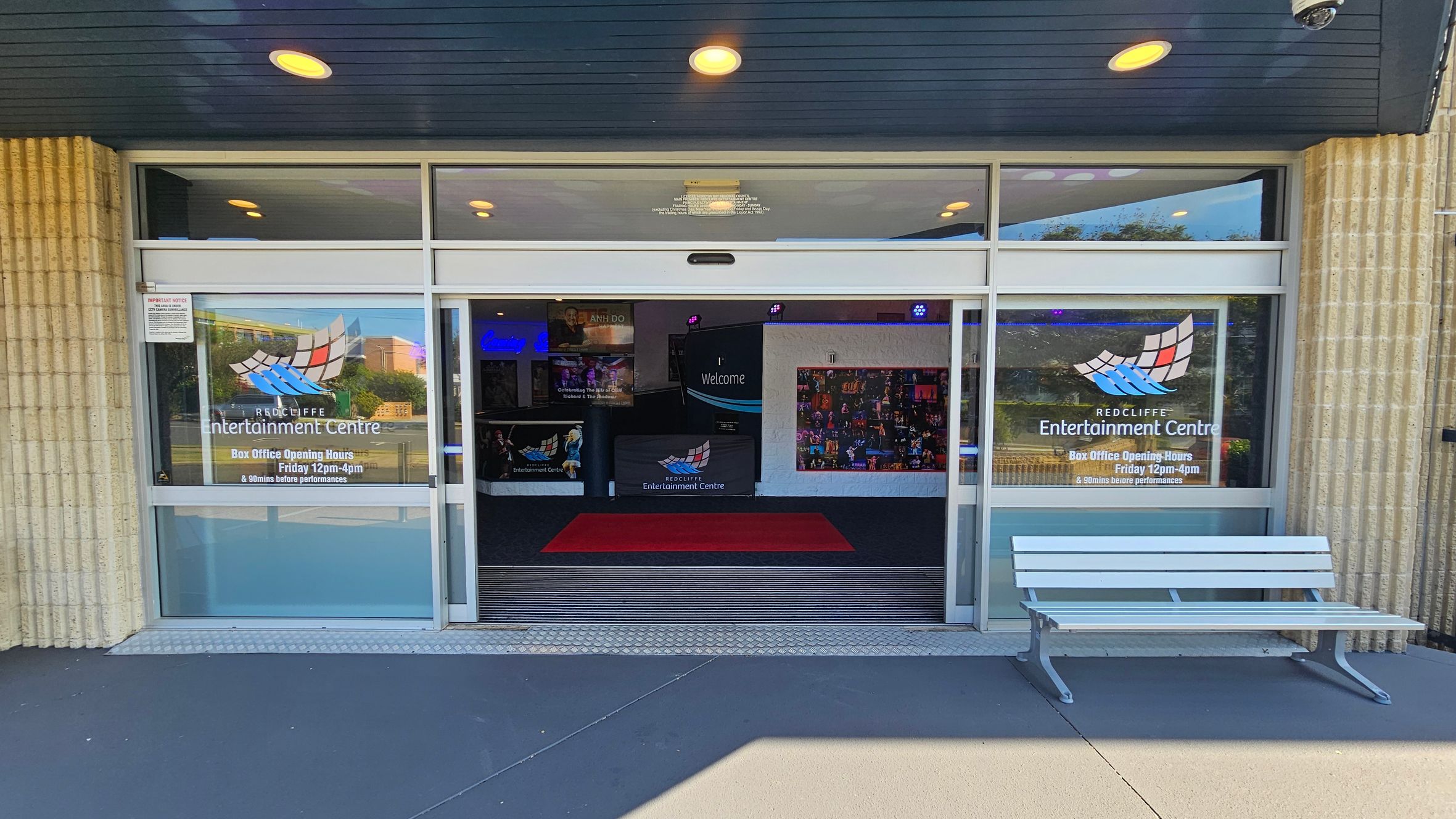 Entrance to a building with large glass doors and windows. The windows display the logo and name "Redcliffe Entertainment Centre" along with box office hours. A white bench is positioned on the right side of the entrance. Inside, a red carpet leads to a welcome sign and colourful posters on the walls. The ceiling is dark with recessed lighting, and the floor outside is grey. Entrance to a building with large glass doors and windows. The windows display the logo and name "Redcliffe Entertainment Centre" along with box office hours. A white bench is positioned on the right side of the entrance. Inside, a red carpet leads to a welcome sign and colourful posters on the walls. The ceiling is dark with recessed lighting, and the floor outside is grey.