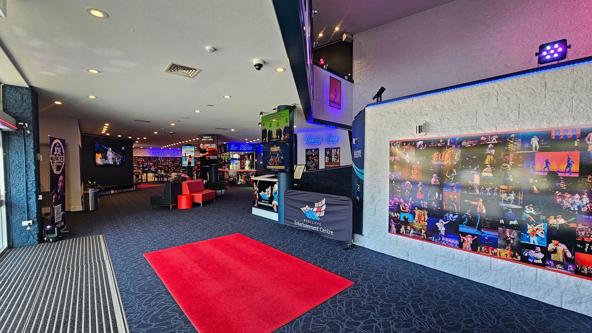 Entrance area with a patterned dark carpet and a red mat leading inside. On the left, a large screen displays a performance, with a poster and a bin nearby. In the centre, a seating area with red and black chairs is visible. The right wall features a collage of performance photos. Above, a sign reads "Coming Soon" with various posters. The ceiling has recessed lighting and a camera. Entrance area with a patterned dark carpet and a red mat leading inside. On the left, a large screen displays a performance, with a poster and a bin nearby. In the centre, a seating area with red and black chairs is visible. The right wall features a collage of performance photos. Above, a sign reads "Coming Soon" with various posters. The ceiling has recessed lighting and a camera.