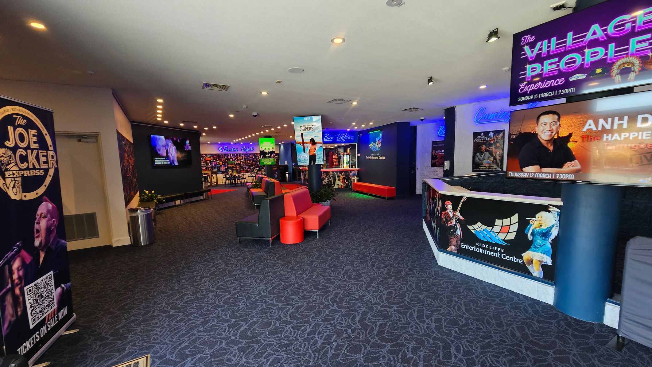 A spacious lobby with a patterned carpet floor. On the left, a large poster for "The Joe Cocker Express" is displayed. In the centre, red and black seating is arranged, with a digital screen showing promotional content. The right side features a counter with posters for upcoming shows, including "The Village People Experience" and "Anh Do: The Happiest." Neon signs for "Theatre" and "Box Office" are visible in the background, along with a wall of colourful lights. The ceiling has recessed lighting. A spacious lobby with a patterned carpet floor. On the left, a large poster for "The Joe Cocker Express" is displayed. In the centre, red and black seating is arranged, with a digital screen showing promotional content. The right side features a counter with posters for upcoming shows, including "The Village People Experience" and "Anh Do: The Happiest." Neon signs for "Theatre" and "Box Office" are visible in the background, along with a wall of colourful lights. The ceiling has recessed lighting.
