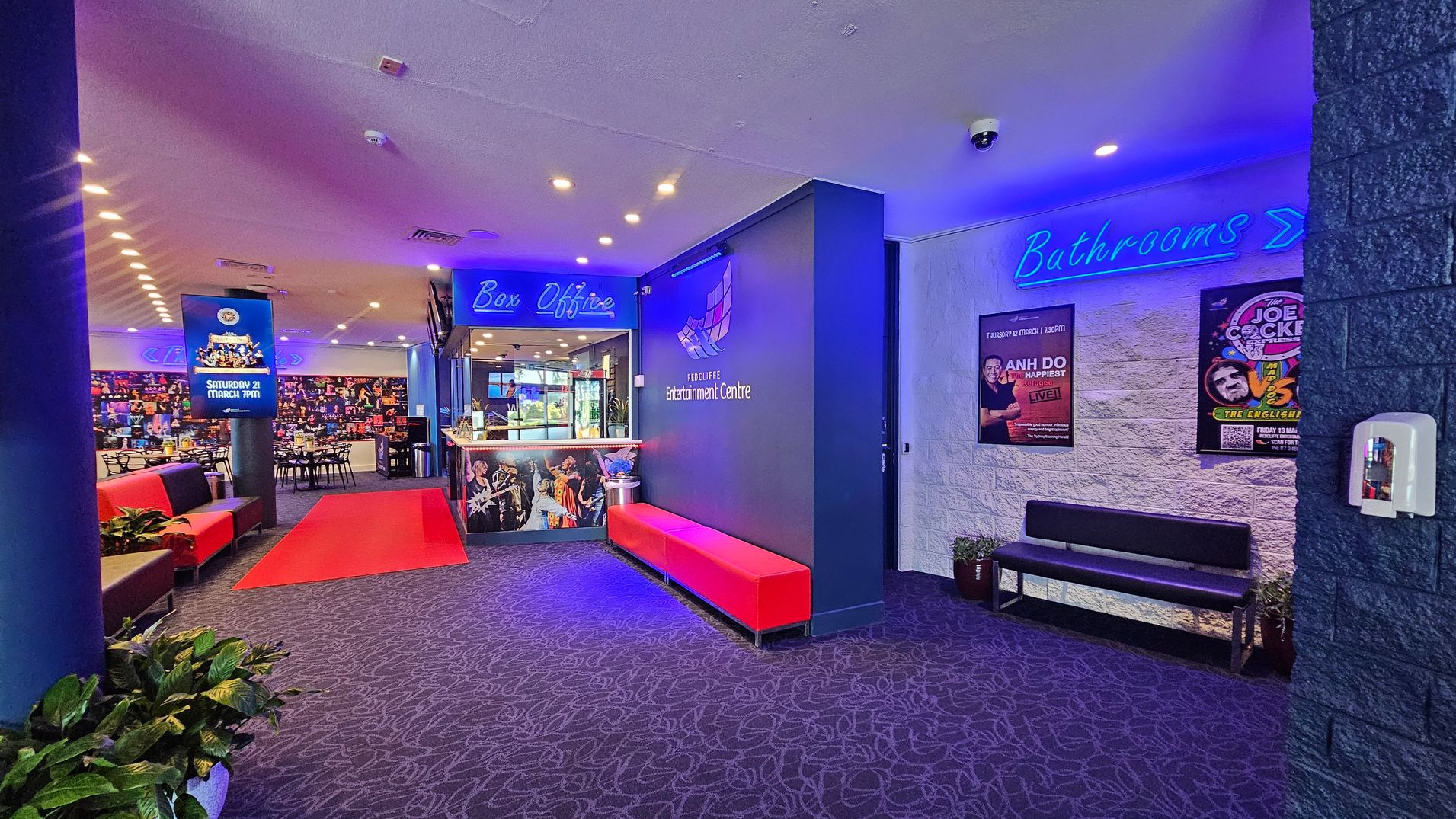 A vibrant entertainment centre lobby with a patterned carpet. On the left, red and black seating lines the wall, with a digital display advertising an event. In the centre, a red carpet leads to the box office, which is illuminated with blue signage. The right wall features posters and a blue neon "Bathrooms" sign above a black bench. Potted plants are placed around the area, and a hand sanitiser dispenser is mounted on the wall. The ceiling has recessed lighting. A vibrant entertainment centre lobby with a patterned carpet. On the left, red and black seating lines the wall, with a digital display advertising an event. In the centre, a red carpet leads to the box office, which is illuminated with blue signage. The right wall features posters and a blue neon "Bathrooms" sign above a black bench. Potted plants are placed around the area, and a hand sanitiser dispenser is mounted on the wall. The ceiling has recessed lighting.
