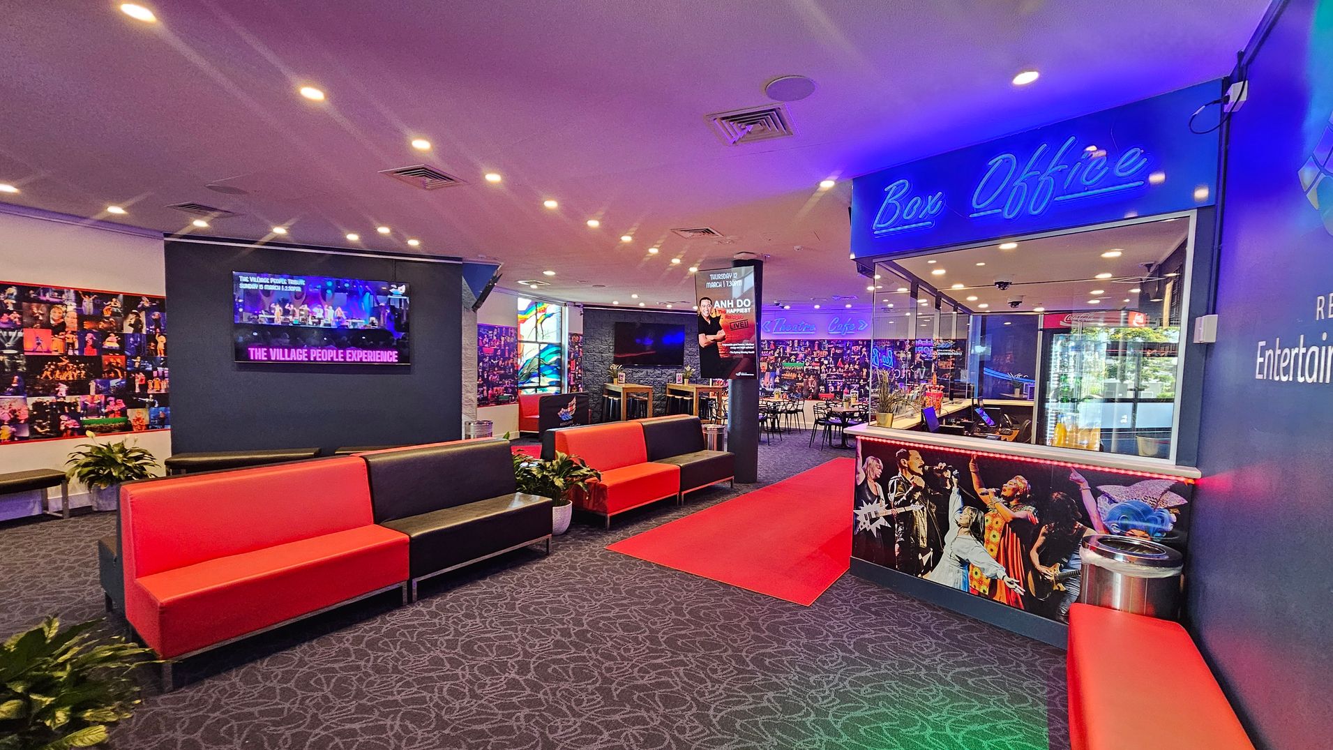 A vibrant lobby area with a patterned carpet and a ceiling dotted with recessed lights. On the left, a wall features a collage of performance images and a screen displaying "The Village People Experience." Red and black cushioned benches are arranged along the wall. On the right, a box office with a neon sign and a counter decorated with performer images. In the background, a café area with tables and chairs is visible, along with a red carpet leading towards it. A vibrant lobby area with a patterned carpet and a ceiling dotted with recessed lights. On the left, a wall features a collage of performance images and a screen displaying "The Village People Experience." Red and black cushioned benches are arranged along the wall. On the right, a box office with a neon sign and a counter decorated with performer images. In the background, a café area with tables and chairs is visible, along with a red carpet leading towards it.