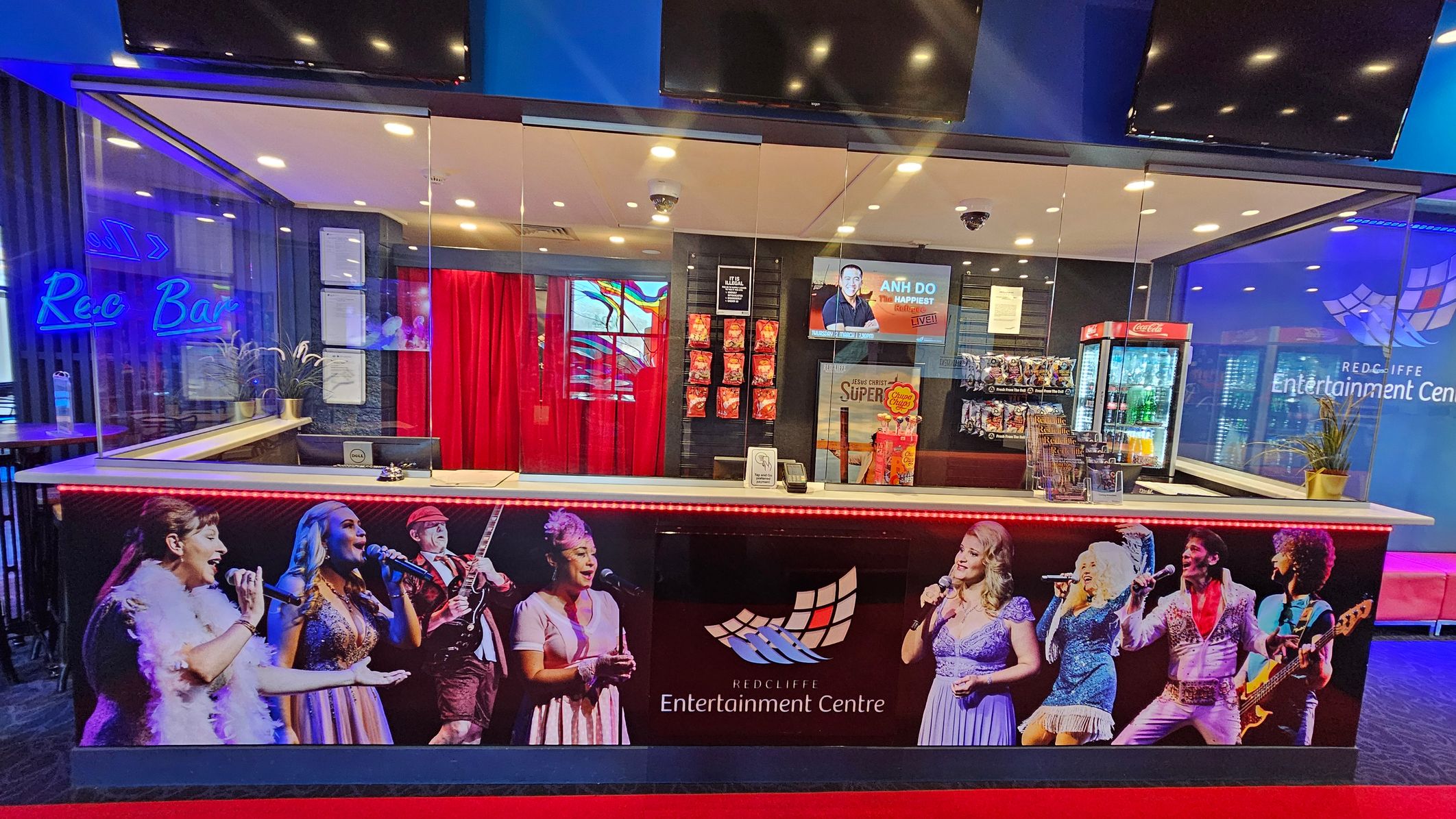A ticket counter at the Redcliffe Entertainment Centre with a glass partition. The counter features images of performers and the centre's logo. Behind the counter, there are red curtains, a display of snacks, and a refrigerator with drinks. A digital screen shows an advertisement. To the left, a neon sign reads "Rec Bar" with seating visible. The floor is carpeted in dark tones, and the ceiling has recessed lighting. A ticket counter at the Redcliffe Entertainment Centre with a glass partition. The counter features images of performers and the centre's logo. Behind the counter, there are red curtains, a display of snacks, and a refrigerator with drinks. A digital screen shows an advertisement. To the left, a neon sign reads "Rec Bar" with seating visible. The floor is carpeted in dark tones, and the ceiling has recessed lighting.