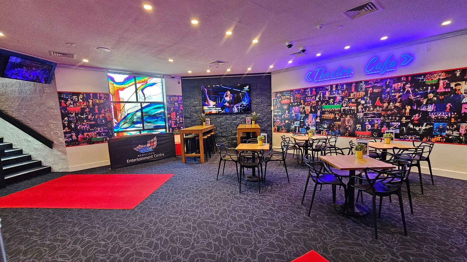 A café area with a patterned carpet and several wooden tables surrounded by black chairs. On the right, a wall is covered with a collage of performance photos and a neon sign reading "Theatre Cafe." In the centre, a large screen displays a performance. To the left, a stained glass window adds colour, and a staircase leads upwards. A red carpet is placed on the floor near the entrance, and a counter with "Redcliffe Entertainment Centre" signage is visible. Ceiling lights illuminate the space. A café area with a patterned carpet and several wooden tables surrounded by black chairs. On the right, a wall is covered with a collage of performance photos and a neon sign reading "Theatre Cafe." In the centre, a large screen displays a performance. To the left, a stained glass window adds colour, and a staircase leads upwards. A red carpet is placed on the floor near the entrance, and a counter with "Redcliffe Entertainment Centre" signage is visible. Ceiling lights illuminate the space.