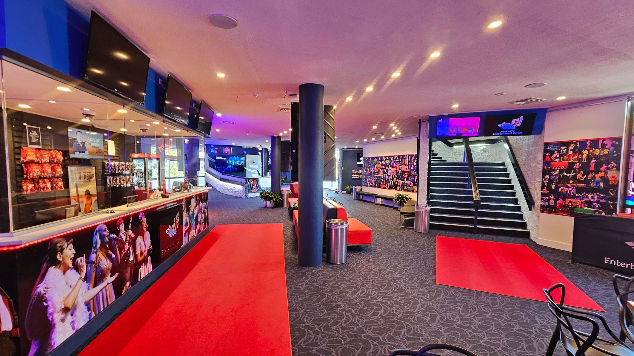A spacious lobby with a red carpet running through the centre. On the left, there's a counter with a glass display, featuring snacks and promotional posters. Above the counter, several screens are mounted. In the middle, a black column stands between red benches and potted plants. On the right, a staircase leads upwards, flanked by photo collages on the walls. The ceiling is dotted with recessed lights, and the floor is covered with a patterned carpet. A spacious lobby with a red carpet running through the centre. On the left, there's a counter with a glass display, featuring snacks and promotional posters. Above the counter, several screens are mounted. In the middle, a black column stands between red benches and potted plants. On the right, a staircase leads upwards, flanked by photo collages on the walls. The ceiling is dotted with recessed lights, and the floor is covered with a patterned carpet.