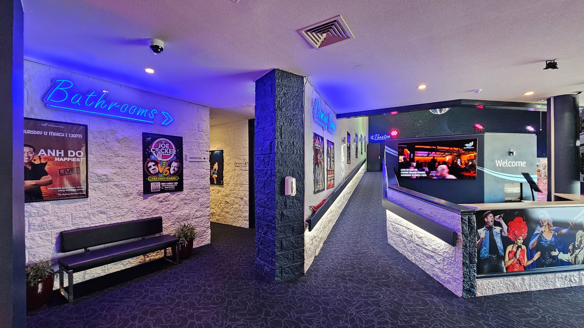 A corridor with a dark patterned carpet and white brick walls. On the left, a blue neon sign reads "Bathrooms" with an arrow pointing right, above a bench and posters. The right side features a ramp leading to a theatre, with a "Welcome" sign and a large screen displaying images. Posters and a neon "Theatre" sign are visible along the wall. The ceiling is light-coloured with a security camera and vents. A corridor with a dark patterned carpet and white brick walls. On the left, a blue neon sign reads "Bathrooms" with an arrow pointing right, above a bench and posters. The right side features a ramp leading to a theatre, with a "Welcome" sign and a large screen displaying images. Posters and a neon "Theatre" sign are visible along the wall. The ceiling is light-coloured with a security camera and vents.