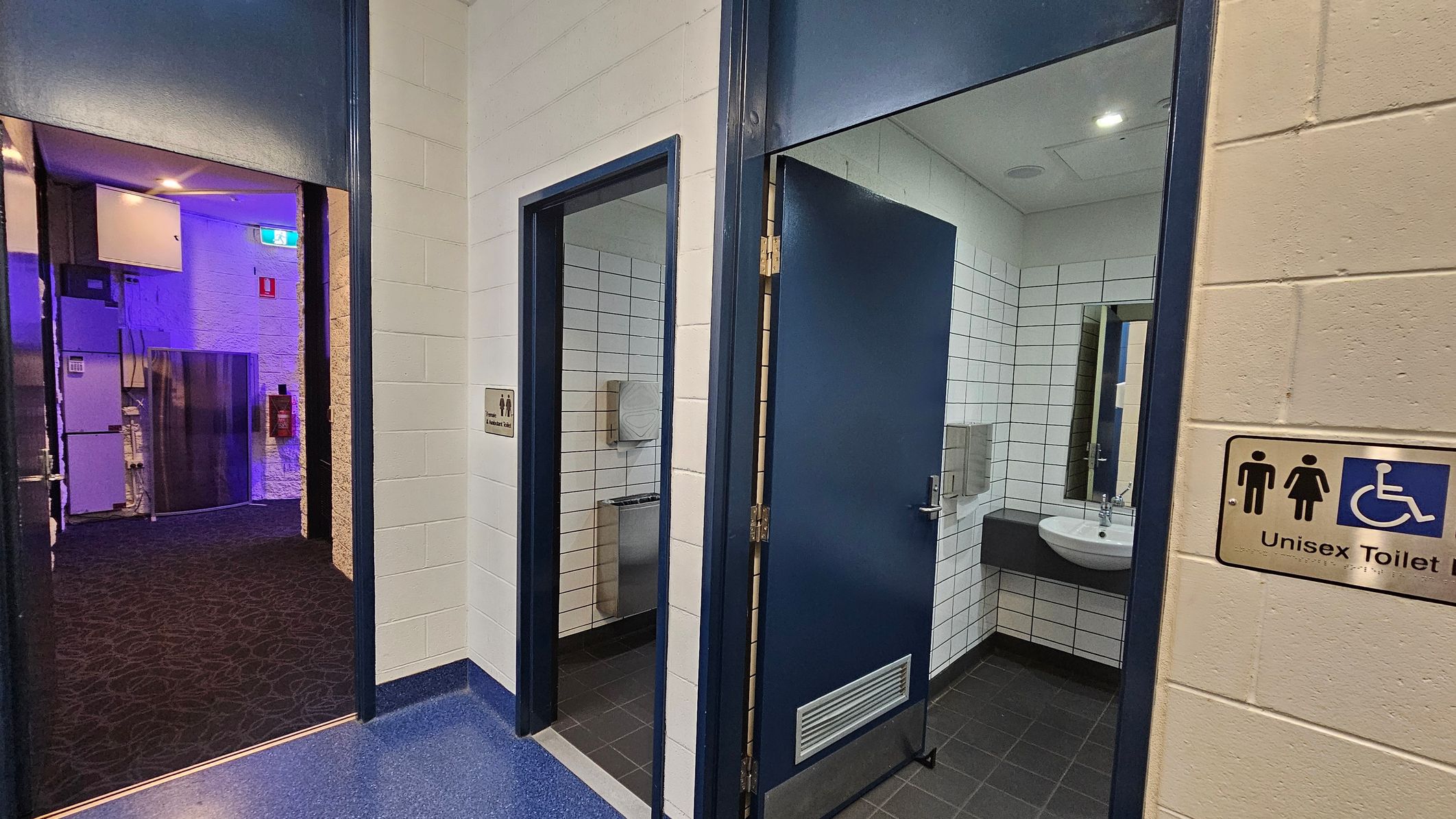 A corridor with two open doors leading to unisex toilets. The left door reveals a toilet with a paper dispenser and a stainless steel fixture. The right door shows a sink with a mirror above it. Both rooms have white tiled walls. The floor is dark, and the ceiling has recessed lighting. A sign on the right wall indicates the toilets are unisex and accessible. The corridor outside has a dark carpet and a blue-lit area with visible electrical panels. A corridor with two open doors leading to unisex toilets. The left door reveals a toilet with a paper dispenser and a stainless steel fixture. The right door shows a sink with a mirror above it. Both rooms have white tiled walls. The floor is dark, and the ceiling has recessed lighting. A sign on the right wall indicates the toilets are unisex and accessible. The corridor outside has a dark carpet and a blue-lit area with visible electrical panels.