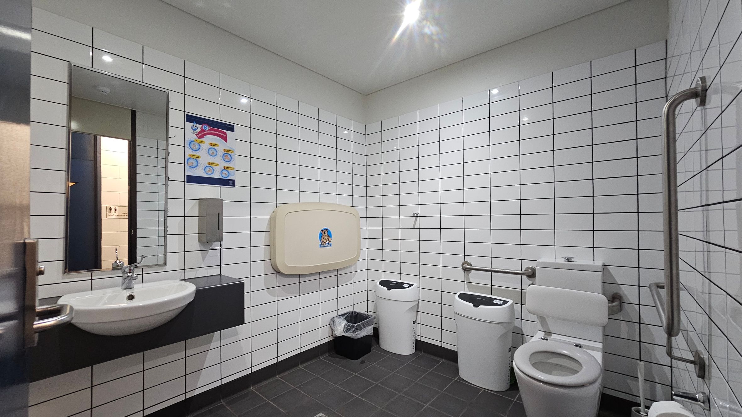 A public restroom with white tiled walls and a dark tiled floor. On the left, a sink with a mirror above it and a soap dispenser nearby. In the centre, a baby changing station is mounted on the wall, with a poster above it. Below, a small bin with a liner. On the right, a toilet with support rails and two sanitary bins. The ceiling is plain with recessed lighting. A public restroom with white tiled walls and a dark tiled floor. On the left, a sink with a mirror above it and a soap dispenser nearby. In the centre, a baby changing station is mounted on the wall, with a poster above it. Below, a small bin with a liner. On the right, a toilet with support rails and two sanitary bins. The ceiling is plain with recessed lighting.