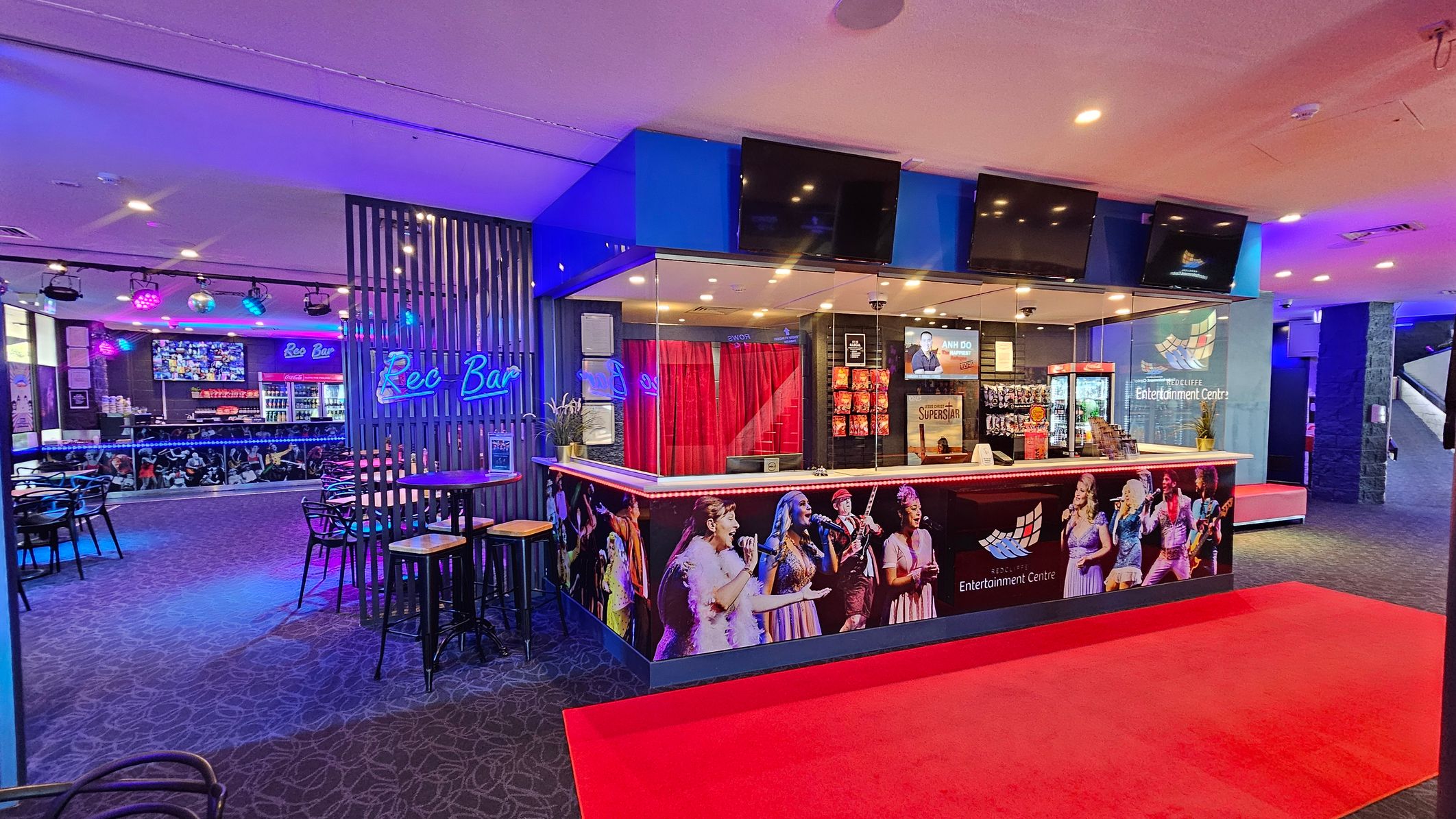 A vibrant entertainment venue with a bar area on the left, featuring a neon "Rec Bar" sign and several tables with stools. The bar counter displays various drinks and a large screen above. In the centre, a reception area with a counter adorned with images of performers. Behind the counter, red curtains and promotional posters are visible. The floor is carpeted with a red carpet leading to the counter, and the ceiling has recessed lighting. A vibrant entertainment venue with a bar area on the left, featuring a neon "Rec Bar" sign and several tables with stools. The bar counter displays various drinks and a large screen above. In the centre, a reception area with a counter adorned with images of performers. Behind the counter, red curtains and promotional posters are visible. The floor is carpeted with a red carpet leading to the counter, and the ceiling has recessed lighting.