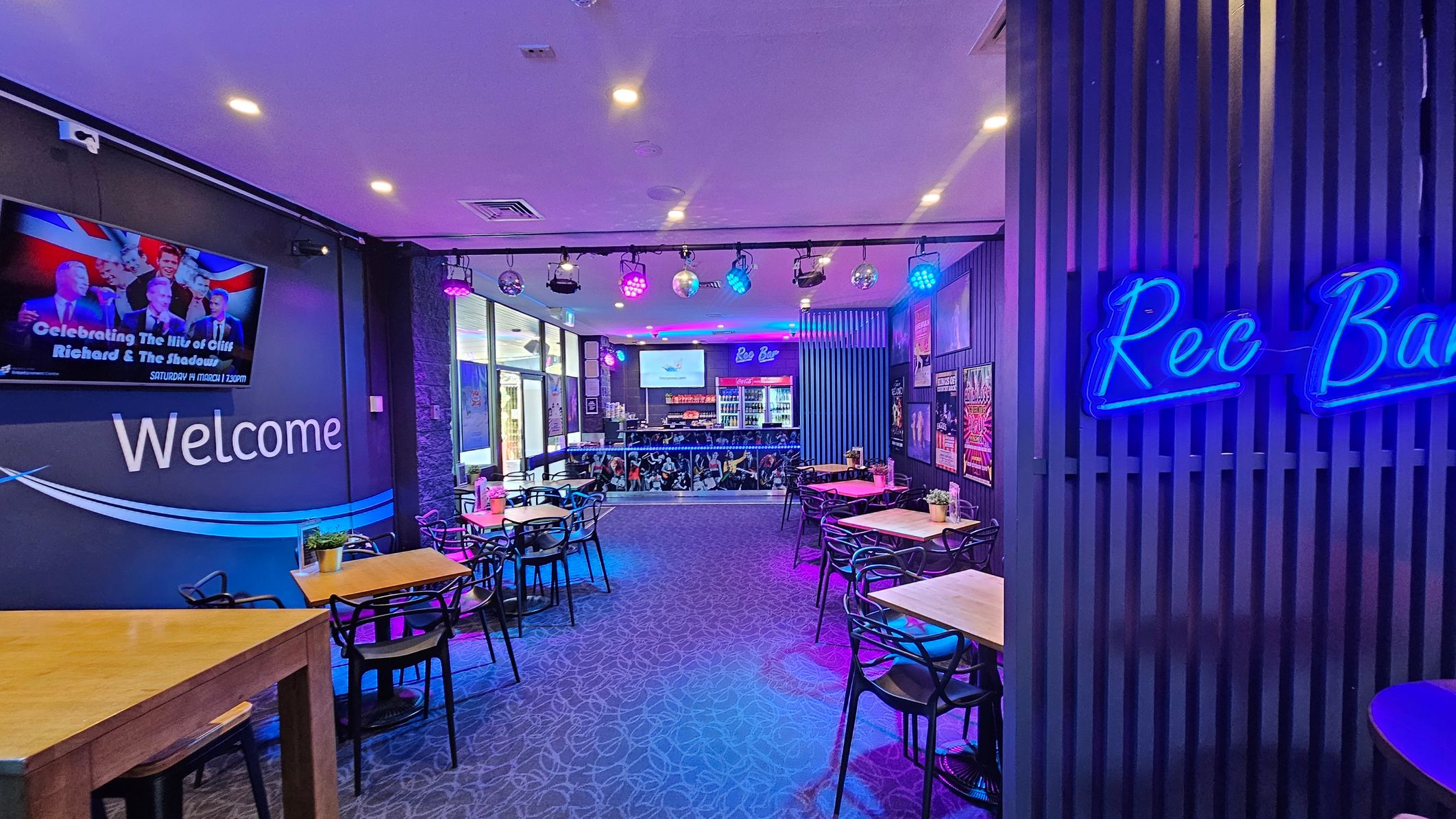 A vibrant bar interior with a purple and blue lighting theme. On the left, a wall-mounted screen displays an event poster above the word "Welcome." Several wooden tables with black chairs are arranged throughout the space. On the right, a neon sign reads "Rec Bar" on a slatted wall. The bar area at the back features a counter with drinks and a screen above. The ceiling has spotlights and disco balls, and the carpet has a subtle pattern. A vibrant bar interior with a purple and blue lighting theme. On the left, a wall-mounted screen displays an event poster above the word "Welcome." Several wooden tables with black chairs are arranged throughout the space. On the right, a neon sign reads "Rec Bar" on a slatted wall. The bar area at the back features a counter with drinks and a screen above. The ceiling has spotlights and disco balls, and the carpet has a subtle pattern.