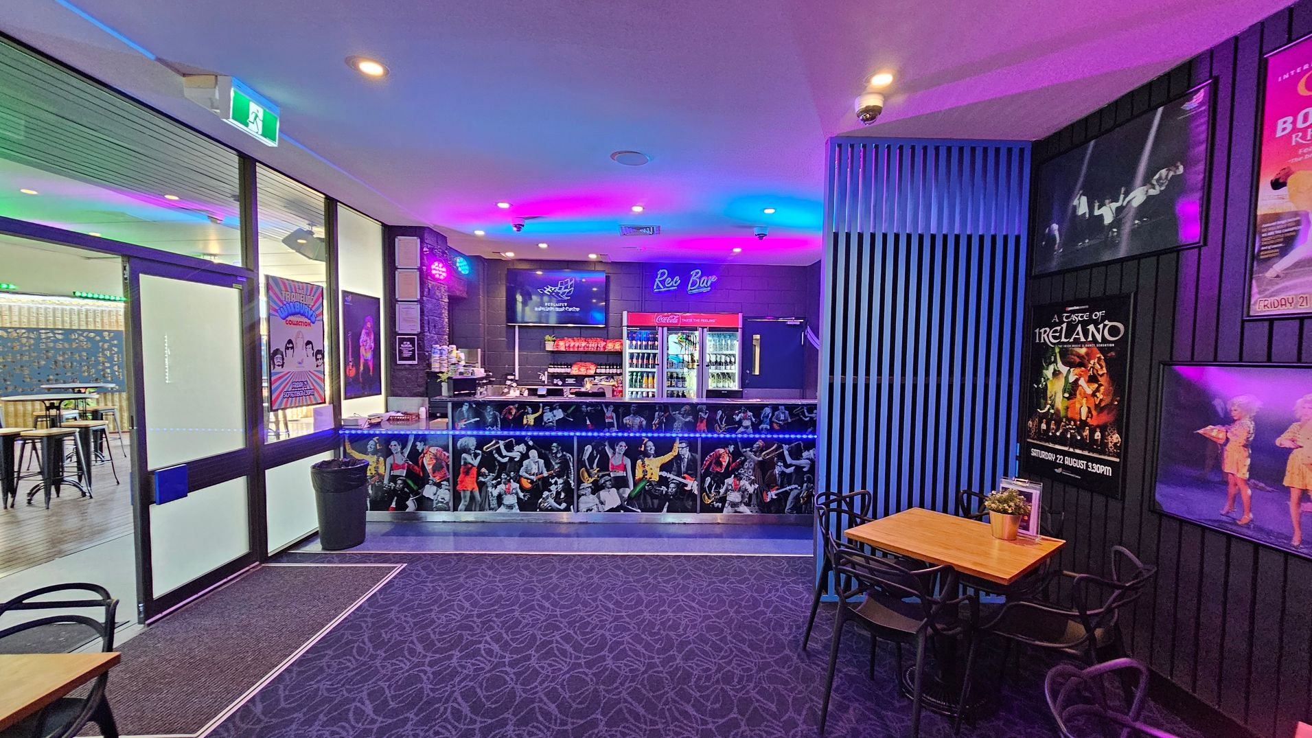A bar area with a vibrant, colourful atmosphere. On the left, there are glass doors with posters and a glimpse of high tables and stools in the adjacent room. The centre features a bar counter decorated with images of musicians, with a neon "Rec Bar" sign above. Behind the counter, there are shelves with drinks and snacks. On the right, a small wooden table with black chairs is set against a wall adorned with various event posters. The ceiling has recessed lighting, and the floor is carpeted with a patterned design. A bar area with a vibrant, colourful atmosphere. On the left, there are glass doors with posters and a glimpse of high tables and stools in the adjacent room. The centre features a bar counter decorated with images of musicians, with a neon "Rec Bar" sign above. Behind the counter, there are shelves with drinks and snacks. On the right, a small wooden table with black chairs is set against a wall adorned with various event posters. The ceiling has recessed lighting, and the floor is carpeted with a patterned design.