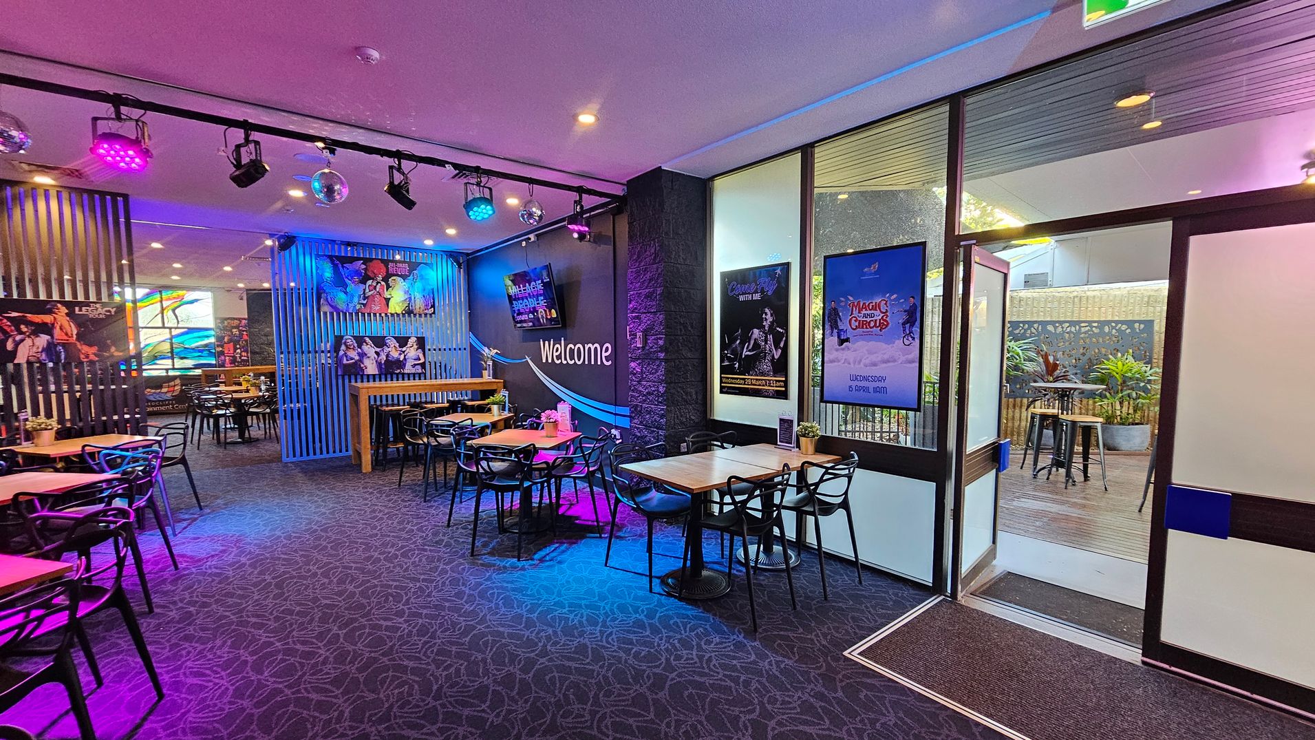 A vibrant indoor space with a patterned carpet floor and a ceiling featuring colourful spotlights and disco balls. On the left, there are tables and chairs, with posters on the walls. In the centre, a "Welcome" sign is visible on a dark wall. To the right, large windows and a glass door lead to an outdoor area with plants and high tables. Posters are displayed on the walls near the entrance. A vibrant indoor space with a patterned carpet floor and a ceiling featuring colourful spotlights and disco balls. On the left, there are tables and chairs, with posters on the walls. In the centre, a "Welcome" sign is visible on a dark wall. To the right, large windows and a glass door lead to an outdoor area with plants and high tables. Posters are displayed on the walls near the entrance.