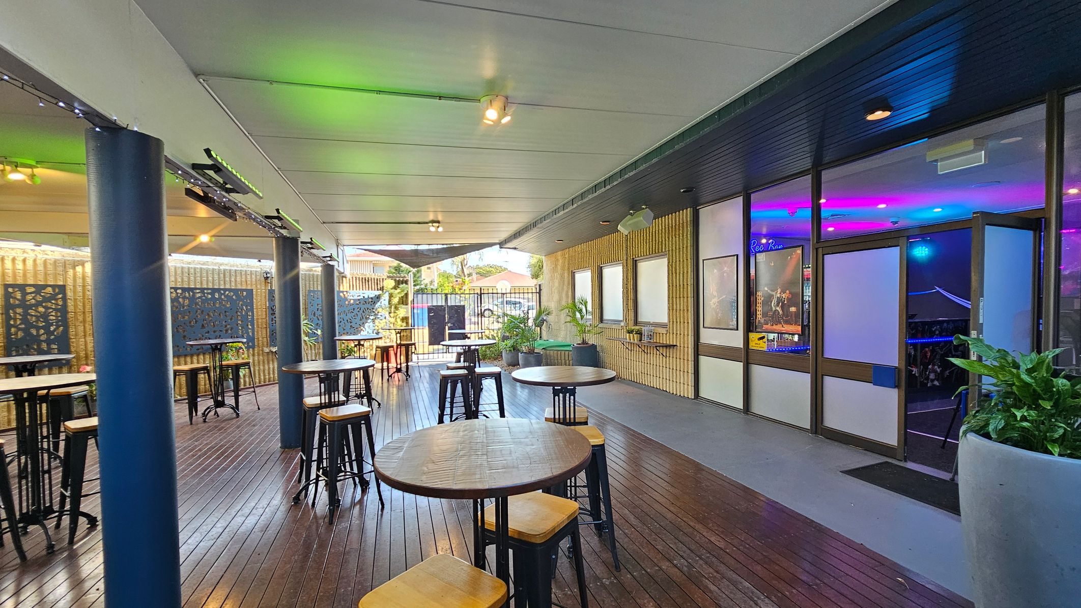 A covered outdoor area with wooden flooring and a white ceiling. On the left, there are several round wooden tables with high stools, and blue decorative panels on the walls. Blue columns support the ceiling. On the right, a glass door leads inside, where colourful lights are visible. Potted plants are placed near the entrance. The background shows a fence and some buildings. A covered outdoor area with wooden flooring and a white ceiling. On the left, there are several round wooden tables with high stools, and blue decorative panels on the walls. Blue columns support the ceiling. On the right, a glass door leads inside, where colourful lights are visible. Potted plants are placed near the entrance. The background shows a fence and some buildings.