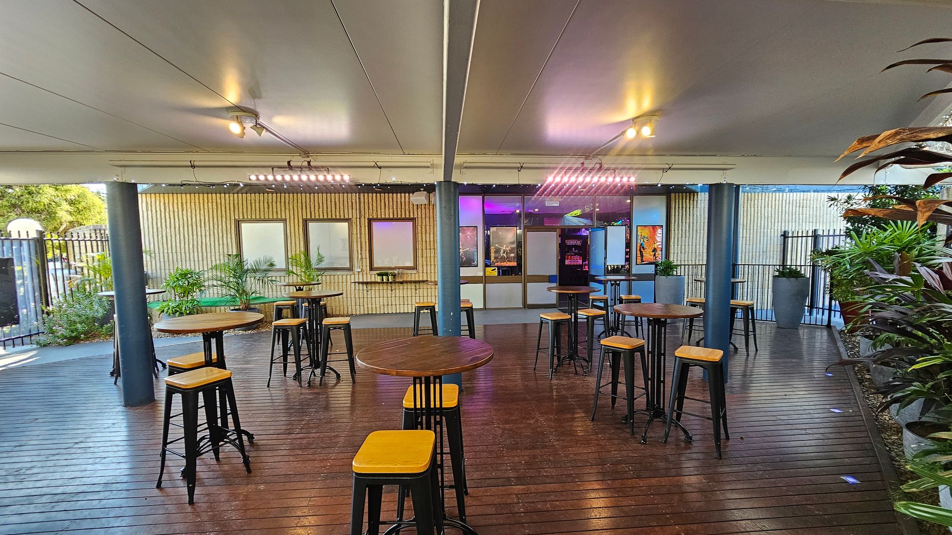 A covered outdoor seating area with wooden flooring and a white ceiling. In the centre, there are several round wooden tables with tall stools. The background features a wall with windows and a door, illuminated by purple and yellow lights. Potted plants are placed along the wall and on the right side. The left side shows a metal fence and more greenery. Ceiling lights provide additional illumination. A covered outdoor seating area with wooden flooring and a white ceiling. In the centre, there are several round wooden tables with tall stools. The background features a wall with windows and a door, illuminated by purple and yellow lights. Potted plants are placed along the wall and on the right side. The left side shows a metal fence and more greenery. Ceiling lights provide additional illumination.