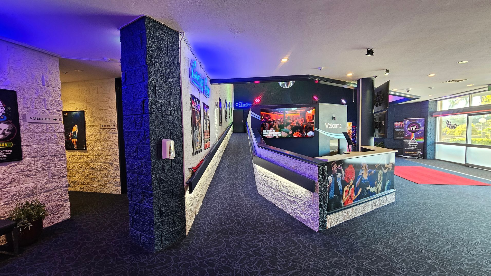 A theatre lobby with a patterned carpeted floor. On the left, a corridor leads to amenities and toilets, with posters on the walls. In the centre, a ramp with more posters leads to a theatre entrance. On the right, a reception desk with a "Welcome" sign and images of performers. The ceiling has recessed lighting, and large windows on the right let in natural light. A red carpet is laid out near the windows. A theatre lobby with a patterned carpeted floor. On the left, a corridor leads to amenities and toilets, with posters on the walls. In the centre, a ramp with more posters leads to a theatre entrance. On the right, a reception desk with a "Welcome" sign and images of performers. The ceiling has recessed lighting, and large windows on the right let in natural light. A red carpet is laid out near the windows.