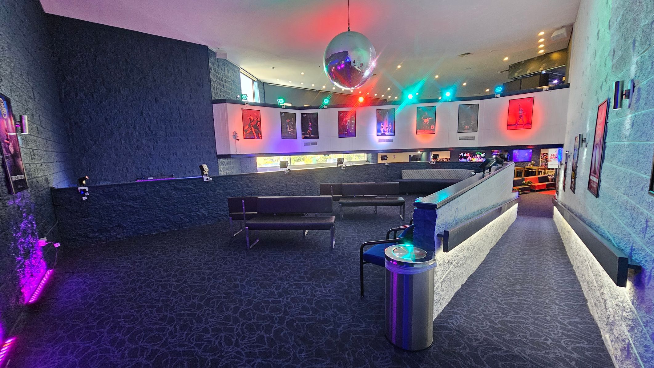 A spacious interior with a dark carpeted floor featuring a swirling pattern. On the left, a textured wall displays several posters, illuminated by purple lighting. In the centre, a large disco ball hangs from the ceiling, casting colourful reflections. Below, black benches are arranged in a seating area. The right side has a sloped walkway with a metal railing and more posters on the wall. The upper level displays additional posters with vibrant lighting effects. A spacious interior with a dark carpeted floor featuring a swirling pattern. On the left, a textured wall displays several posters, illuminated by purple lighting. In the centre, a large disco ball hangs from the ceiling, casting colourful reflections. Below, black benches are arranged in a seating area. The right side has a sloped walkway with a metal railing and more posters on the wall. The upper level displays additional posters with vibrant lighting effects.