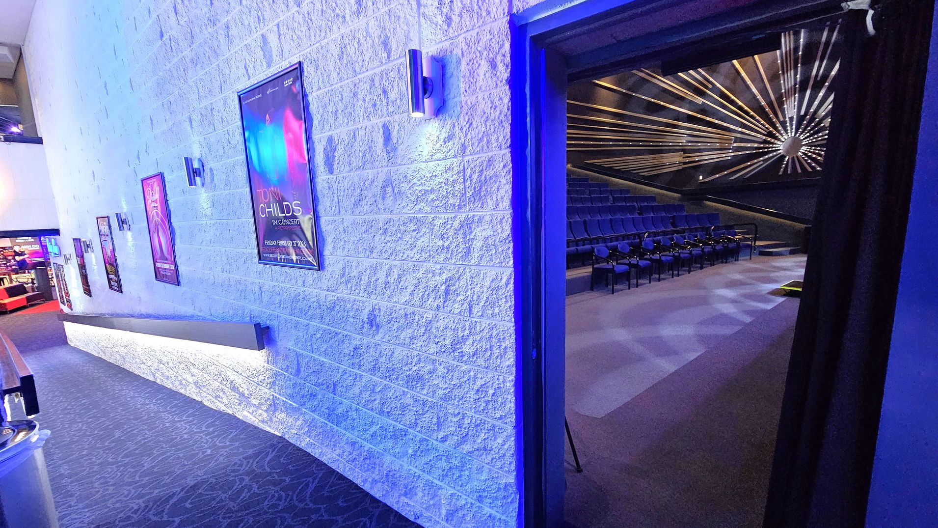 A hallway with a textured white wall on the left, adorned with several posters and modern wall lights. The floor is carpeted with a patterned design. On the right, an open doorway leads into a theatre with rows of blue seats and a striking ceiling design featuring radiating lines and a central circular element. The theatre is dimly lit, contrasting with the brighter hallway. A hallway with a textured white wall on the left, adorned with several posters and modern wall lights. The floor is carpeted with a patterned design. On the right, an open doorway leads into a theatre with rows of blue seats and a striking ceiling design featuring radiating lines and a central circular element. The theatre is dimly lit, contrasting with the brighter hallway.