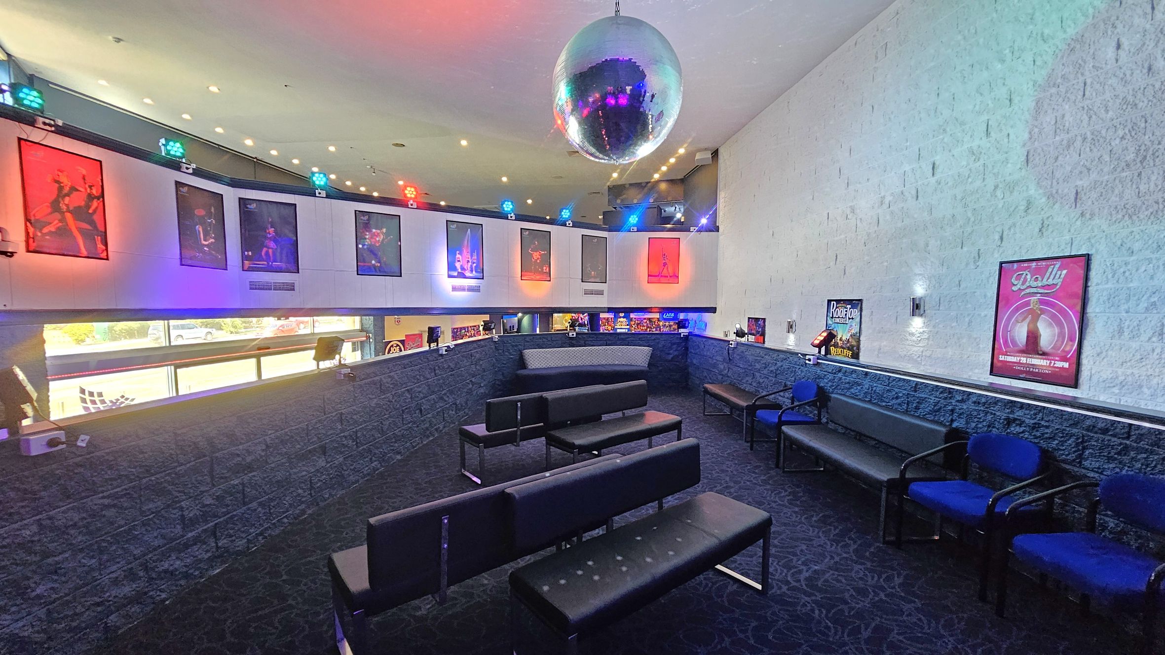A room with a dark carpeted floor and a white ceiling featuring a large disco ball. On the left, there are colourful posters and lights on the upper wall. The seating area consists of black benches and blue chairs arranged in rows. The right wall is adorned with posters, including one for "Dolly." The lower part of the walls is made of dark stone, and there are windows providing a view of the outside. A room with a dark carpeted floor and a white ceiling featuring a large disco ball. On the left, there are colourful posters and lights on the upper wall. The seating area consists of black benches and blue chairs arranged in rows. The right wall is adorned with posters, including one for "Dolly." The lower part of the walls is made of dark stone, and there are windows providing a view of the outside.