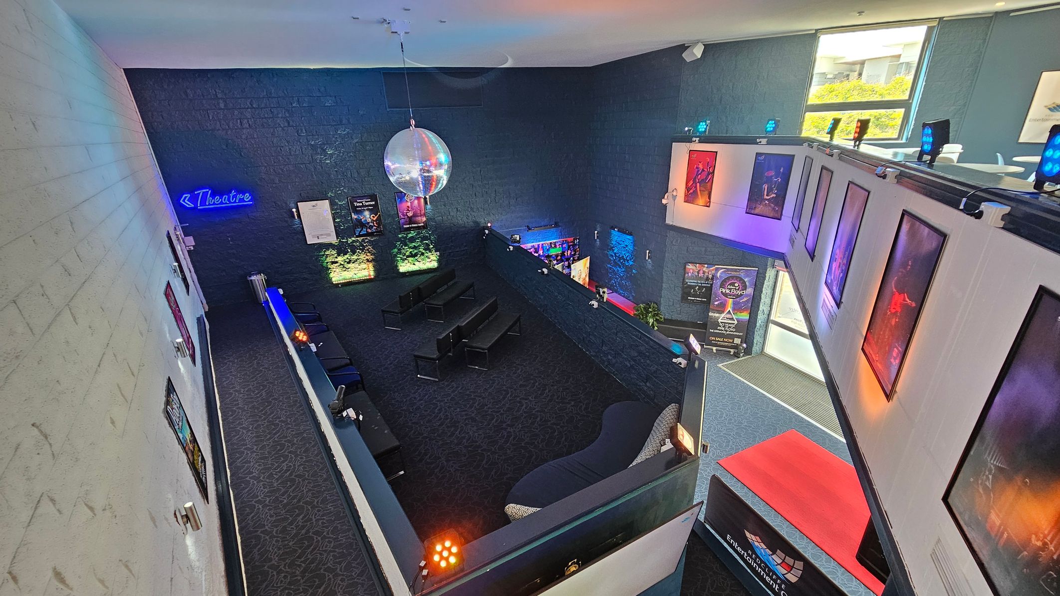 A theatre lobby with a dark carpeted floor and high ceiling. On the left, a white brick wall displays several posters. In the centre, a seating area with black chairs and a large disco ball hanging above. The right side features a balcony with more posters and blue spotlights. A neon sign reading "Theatre" is on the back wall. Bright natural light enters through windows on the upper right, illuminating the space. A theatre lobby with a dark carpeted floor and high ceiling. On the left, a white brick wall displays several posters. In the centre, a seating area with black chairs and a large disco ball hanging above. The right side features a balcony with more posters and blue spotlights. A neon sign reading "Theatre" is on the back wall. Bright natural light enters through windows on the upper right, illuminating the space.