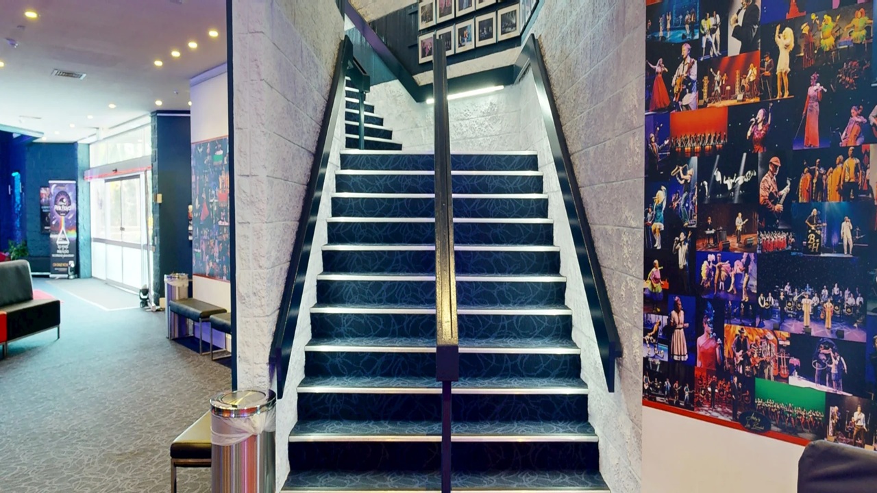 A staircase with dark blue carpet and white walls, featuring a central handrail, leads upwards. On the right, a wall is covered with vibrant performance photos. To the left, a seating area with a dark sofa and a small table is visible. The floor is carpeted in a similar dark tone. The ceiling has recessed lighting, and a large windowed entrance is in the background, allowing natural light to enter. A staircase with dark blue carpet and white walls, featuring a central handrail, leads upwards. On the right, a wall is covered with vibrant performance photos. To the left, a seating area with a dark sofa and a small table is visible. The floor is carpeted in a similar dark tone. The ceiling has recessed lighting, and a large windowed entrance is in the background, allowing natural light to enter.