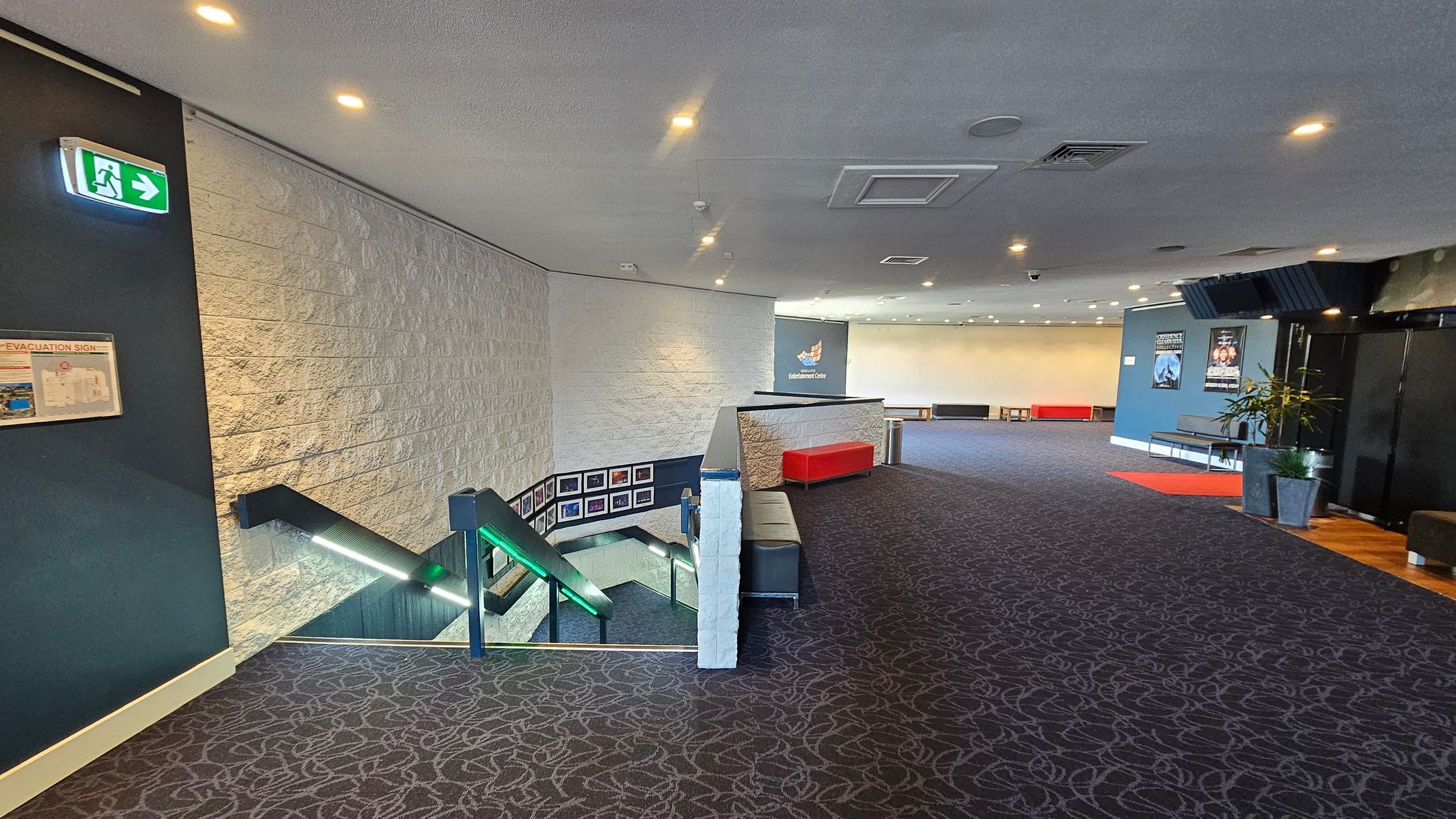 A spacious interior with a patterned carpeted floor. On the left, a staircase with illuminated handrails descends, and a green exit sign is visible above. The wall is textured with a light colour. In the centre, a red bench sits against a low wall. The right side features a blue wall with posters and a potted plant. The ceiling is fitted with recessed lighting and air vents. A spacious interior with a patterned carpeted floor. On the left, a staircase with illuminated handrails descends, and a green exit sign is visible above. The wall is textured with a light colour. In the centre, a red bench sits against a low wall. The right side features a blue wall with posters and a potted plant. The ceiling is fitted with recessed lighting and air vents.