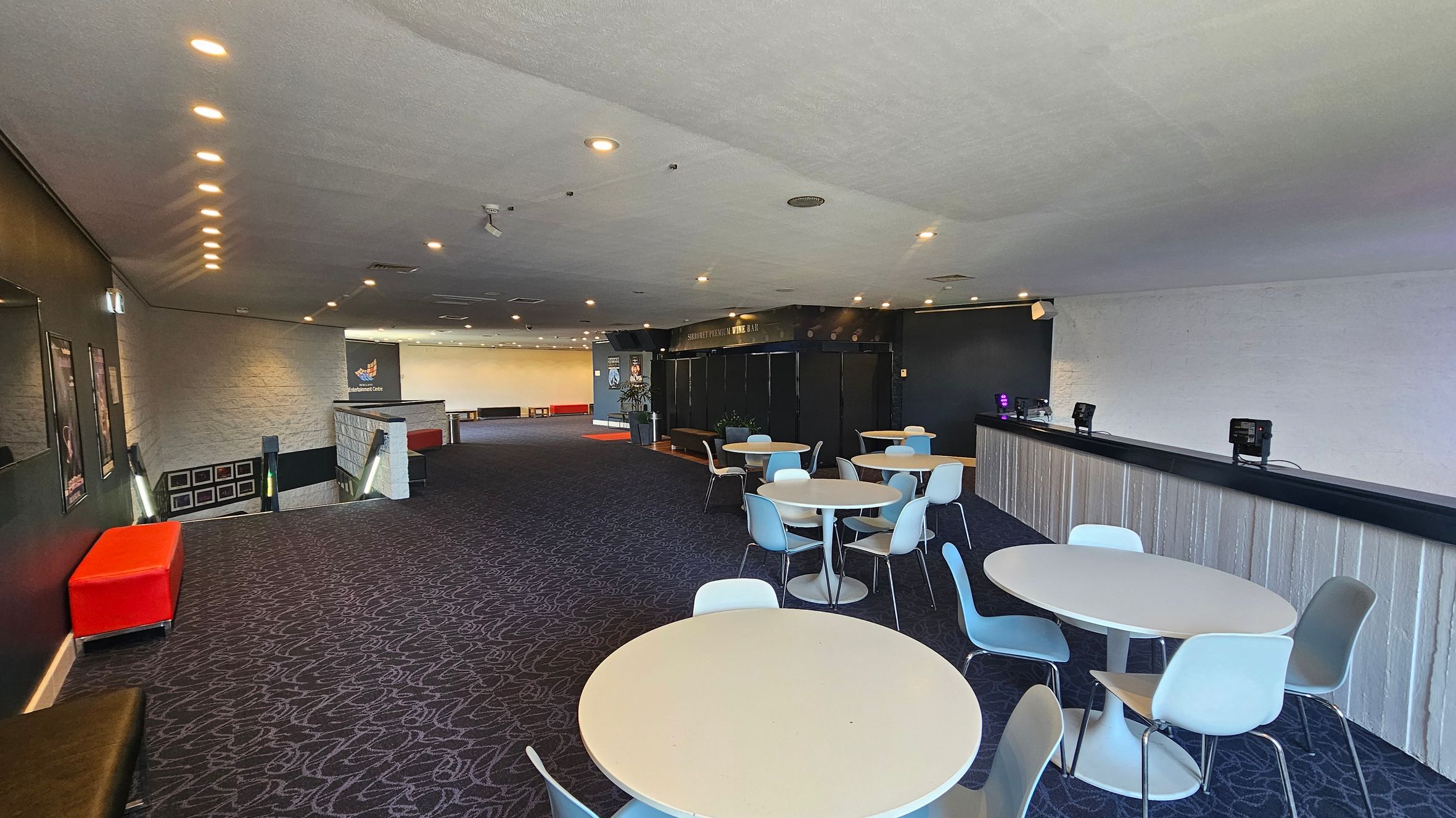A spacious room with a dark patterned carpet and a white ceiling with recessed lighting. On the right, there is a long bar counter with electronic devices on top. In the centre, several round tables with light blue and white chairs are arranged. On the left, a red bench sits near a staircase leading downwards, with framed pictures on the wall. The back wall features a sign and more seating. A spacious room with a dark patterned carpet and a white ceiling with recessed lighting. On the right, there is a long bar counter with electronic devices on top. In the centre, several round tables with light blue and white chairs are arranged. On the left, a red bench sits near a staircase leading downwards, with framed pictures on the wall. The back wall features a sign and more seating.