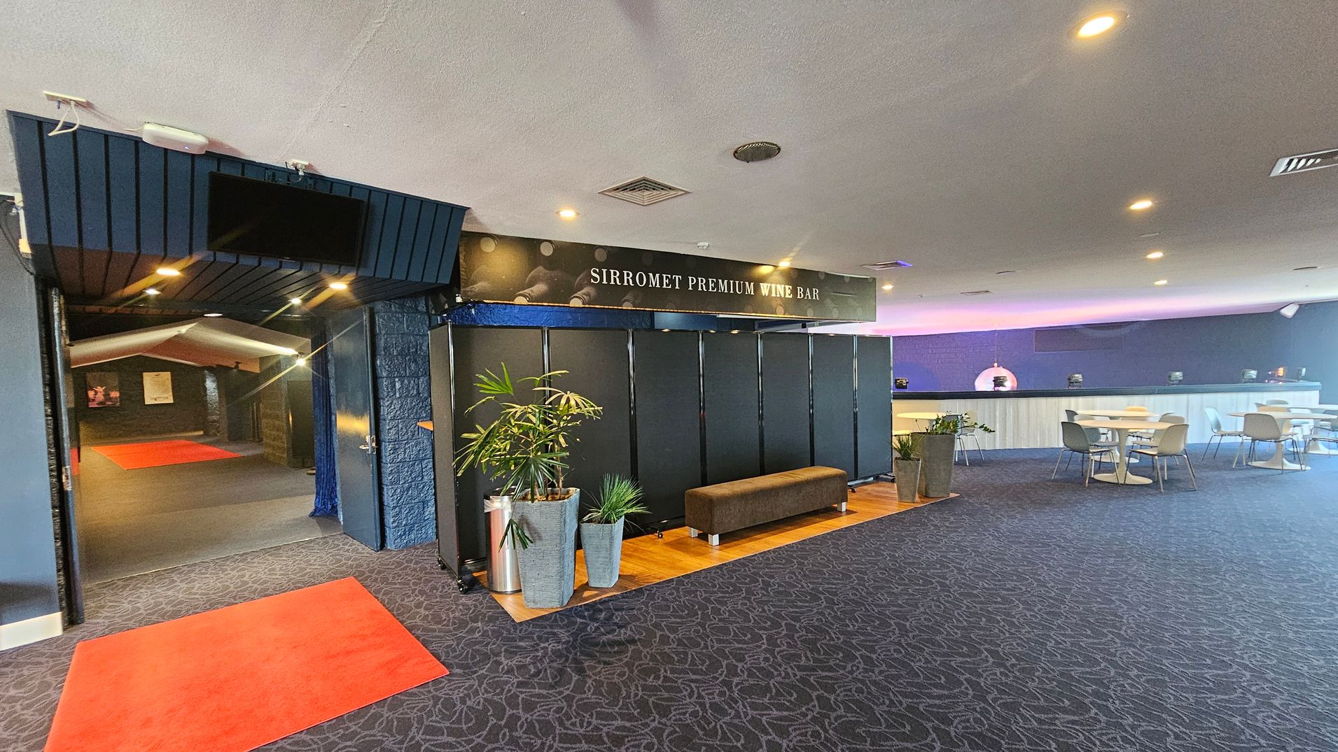 Entrance to a wine bar with a sign reading "Sirromet Premium Wine Bar" above. On the left, a hallway with red carpets leads further inside. In the centre, a seating area with a brown bench and potted plants on a wooden floor. On the right, a spacious area with tables and chairs, and a long counter against the wall. The ceiling is white with recessed lighting, and the floor is covered with patterned carpet. Entrance to a wine bar with a sign reading "Sirromet Premium Wine Bar" above. On the left, a hallway with red carpets leads further inside. In the centre, a seating area with a brown bench and potted plants on a wooden floor. On the right, a spacious area with tables and chairs, and a long counter against the wall. The ceiling is white with recessed lighting, and the floor is covered with patterned carpet.