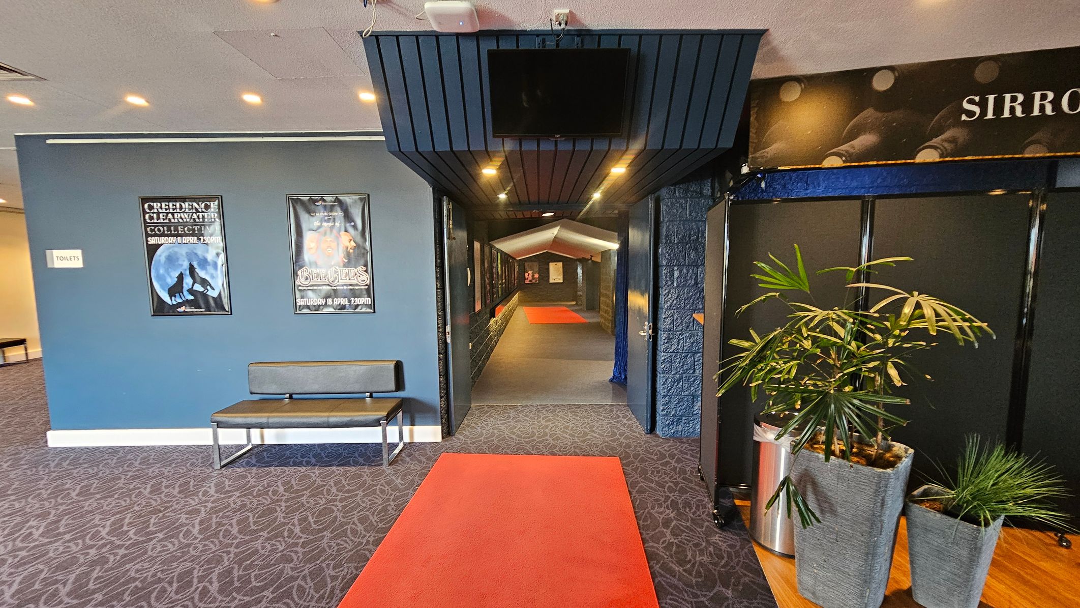 A hallway with a red carpet leading into a corridor. On the left, a blue wall features two posters and a sign for toilets, with a bench below. On the right, there are two potted plants on a wooden surface. The ceiling has recessed lighting, and a black panel with a screen is above the entrance to the corridor. The floor is covered with a patterned carpet. A hallway with a red carpet leading into a corridor. On the left, a blue wall features two posters and a sign for toilets, with a bench below. On the right, there are two potted plants on a wooden surface. The ceiling has recessed lighting, and a black panel with a screen is above the entrance to the corridor. The floor is covered with a patterned carpet.