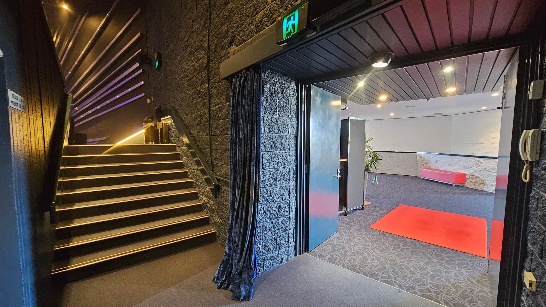 On the left, a staircase with illuminated steps ascends alongside a textured wall. Above the stairs, a series of horizontal lights create a pattern. On the right, an open door leads to a room with a red carpet on a patterned floor. The room has a red bench against a light-coloured wall and a potted plant. The ceiling is dark with recessed lighting. An emergency exit sign is above the door. A curtain hangs beside the door. On the left, a staircase with illuminated steps ascends alongside a textured wall. Above the stairs, a series of horizontal lights create a pattern. On the right, an open door leads to a room with a red carpet on a patterned floor. The room has a red bench against a light-coloured wall and a potted plant. The ceiling is dark with recessed lighting. An emergency exit sign is above the door. A curtain hangs beside the door.
