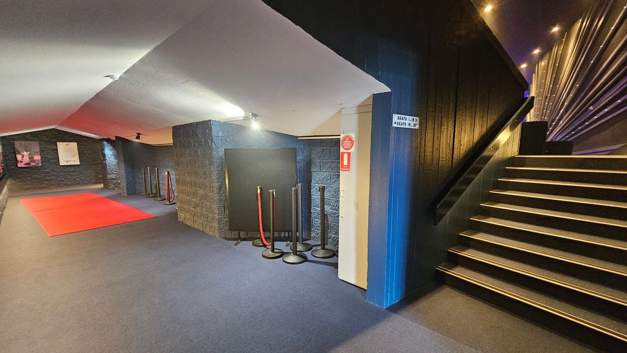 A dimly lit interior with a red carpet on the left side of the floor, leading towards a dark stone wall with two framed posters. In the centre, there are several stanchions with red ropes. On the right, a staircase with illuminated steps ascends alongside a textured wall with vertical lines. The ceiling is slanted and white, with spotlights providing soft lighting. A sign on the wall indicates seating directions. A dimly lit interior with a red carpet on the left side of the floor, leading towards a dark stone wall with two framed posters. In the centre, there are several stanchions with red ropes. On the right, a staircase with illuminated steps ascends alongside a textured wall with vertical lines. The ceiling is slanted and white, with spotlights providing soft lighting. A sign on the wall indicates seating directions.