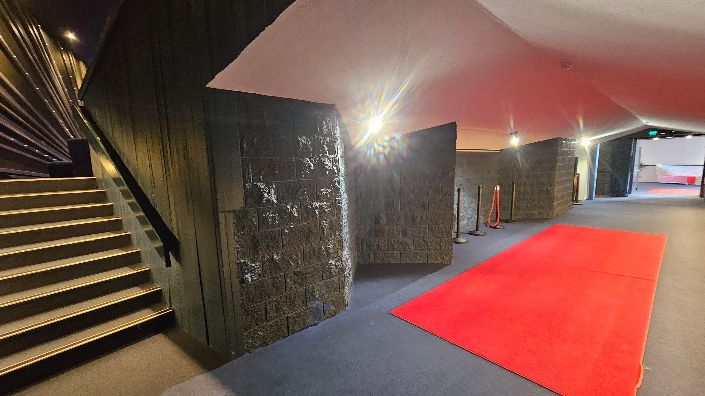 A hallway with a red carpet on the right side, leading towards an open area in the distance. The floor is covered with a dark carpet. On the left, there is a staircase with golden-edged steps and a black handrail. The walls are textured and dark, with bright lights illuminating the space. The ceiling is angled and light-coloured. There are stanchions with red ropes near the walls. A hallway with a red carpet on the right side, leading towards an open area in the distance. The floor is covered with a dark carpet. On the left, there is a staircase with golden-edged steps and a black handrail. The walls are textured and dark, with bright lights illuminating the space. The ceiling is angled and light-coloured. There are stanchions with red ropes near the walls.