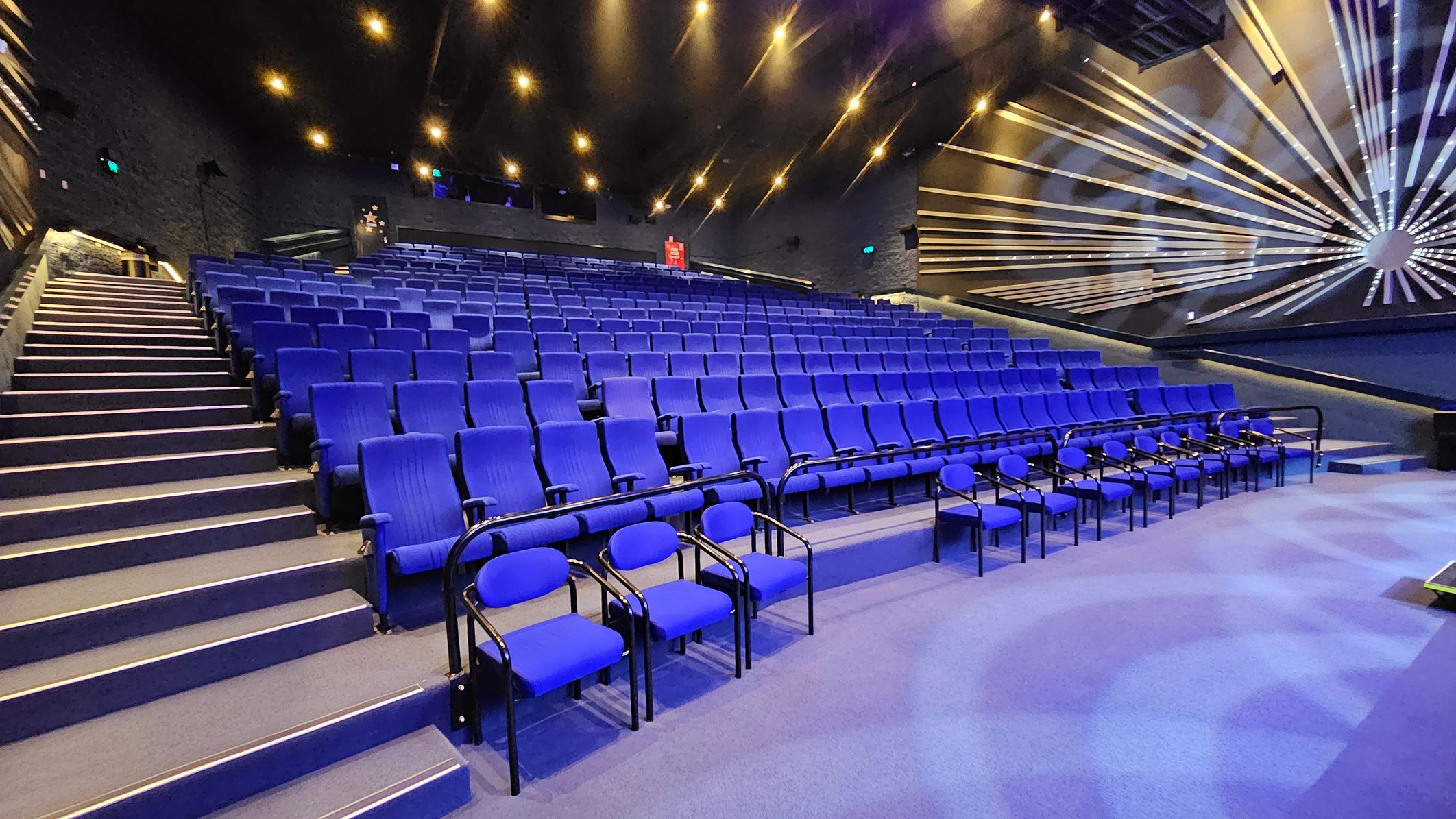 A theatre auditorium with rows of blue seats arranged in tiers. On the left, a staircase with handrails leads up alongside the seating. The right side features a wall with a decorative pattern of radiating lines. The ceiling is dark with numerous small lights, creating a starry effect. The floor is carpeted in blue, matching the seats. A theatre auditorium with rows of blue seats arranged in tiers. On the left, a staircase with handrails leads up alongside the seating. The right side features a wall with a decorative pattern of radiating lines. The ceiling is dark with numerous small lights, creating a starry effect. The floor is carpeted in blue, matching the seats.