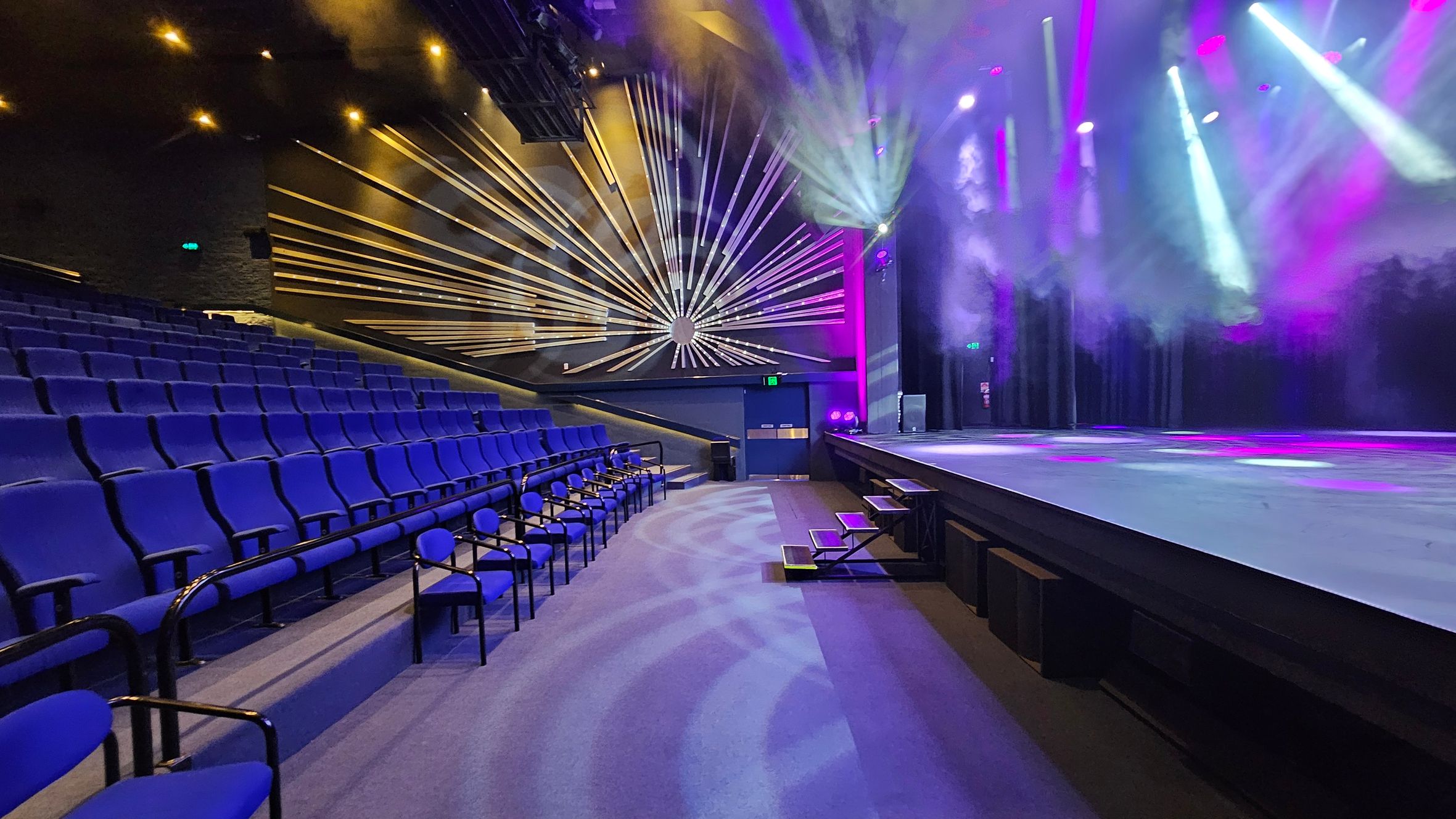 A theatre interior with rows of blue seats on the left, leading up to a stage on the right. The stage is illuminated with colourful lights, casting beams and smoke effects. The back wall features a decorative design with radiating lines. The floor is carpeted, and there are small steps leading up to the stage. The ceiling has spotlights and lighting equipment. A theatre interior with rows of blue seats on the left, leading up to a stage on the right. The stage is illuminated with colourful lights, casting beams and smoke effects. The back wall features a decorative design with radiating lines. The floor is carpeted, and there are small steps leading up to the stage. The ceiling has spotlights and lighting equipment.
