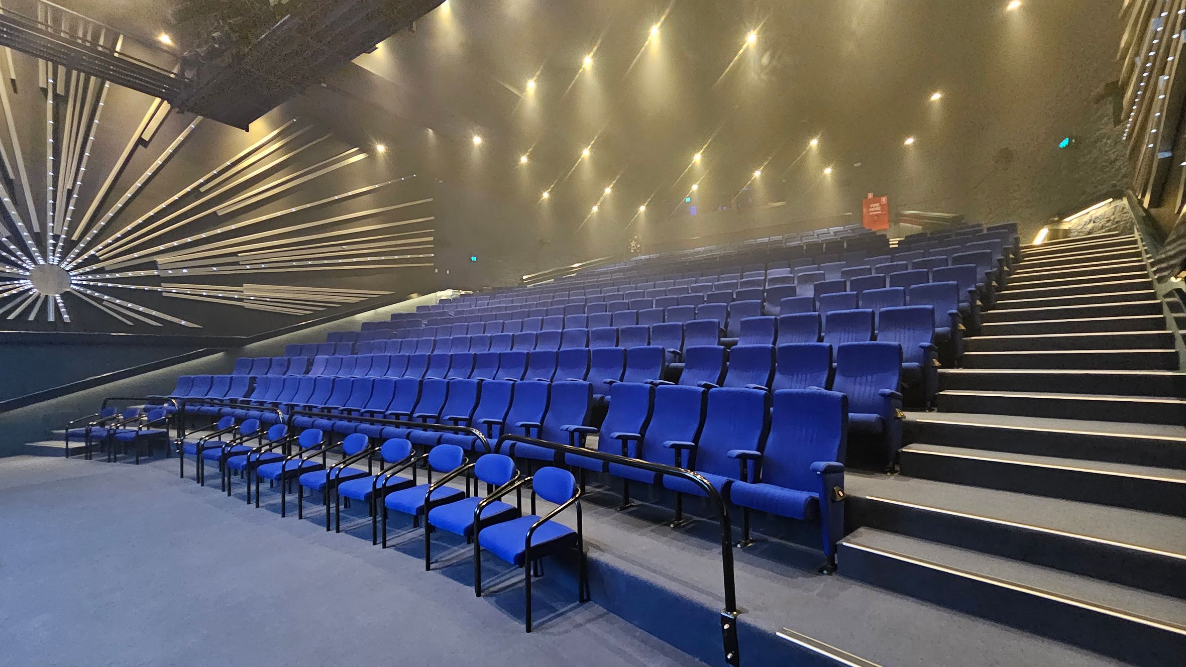 A theatre auditorium with rows of blue seats arranged in a tiered fashion. On the left, there is a striking wall design featuring a radial pattern of metallic lines emanating from a central point. The right side shows a staircase with illuminated steps leading up the seating area. The ceiling is adorned with numerous small spotlights, creating a well-lit atmosphere. The floor is carpeted in a matching blue tone. A theatre auditorium with rows of blue seats arranged in a tiered fashion. On the left, there is a striking wall design featuring a radial pattern of metallic lines emanating from a central point. The right side shows a staircase with illuminated steps leading up the seating area. The ceiling is adorned with numerous small spotlights, creating a well-lit atmosphere. The floor is carpeted in a matching blue tone.