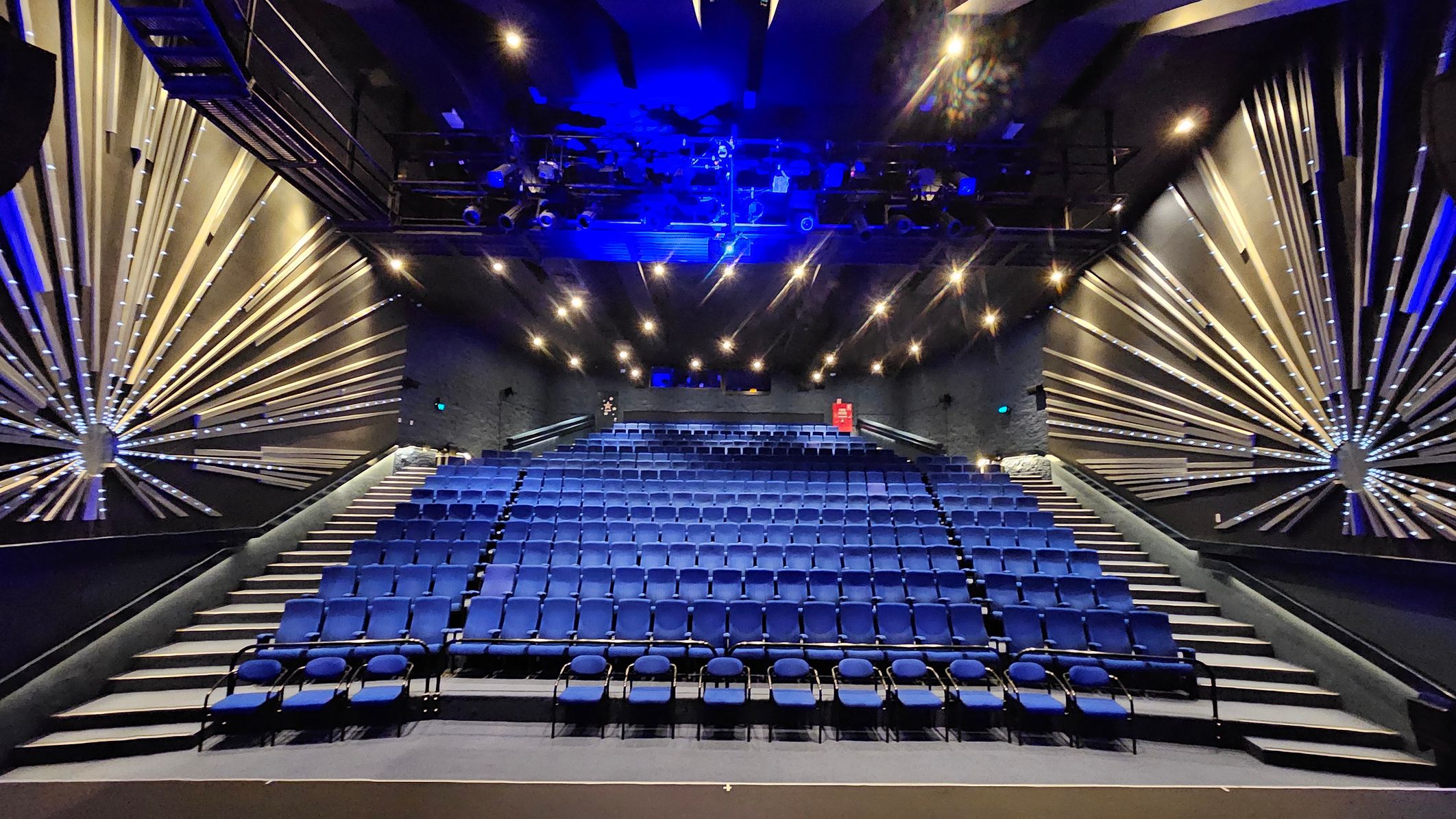 A theatre interior with rows of blue seats arranged in tiers. The walls on both sides feature decorative radial patterns with illuminated lines extending from circular centres. The ceiling is equipped with numerous spotlights and stage lighting, casting a blue hue. The floor is carpeted, and there are aisles on either side of the seating area. A red fire extinguisher is visible on the back wall. A theatre interior with rows of blue seats arranged in tiers. The walls on both sides feature decorative radial patterns with illuminated lines extending from circular centres. The ceiling is equipped with numerous spotlights and stage lighting, casting a blue hue. The floor is carpeted, and there are aisles on either side of the seating area. A red fire extinguisher is visible on the back wall.