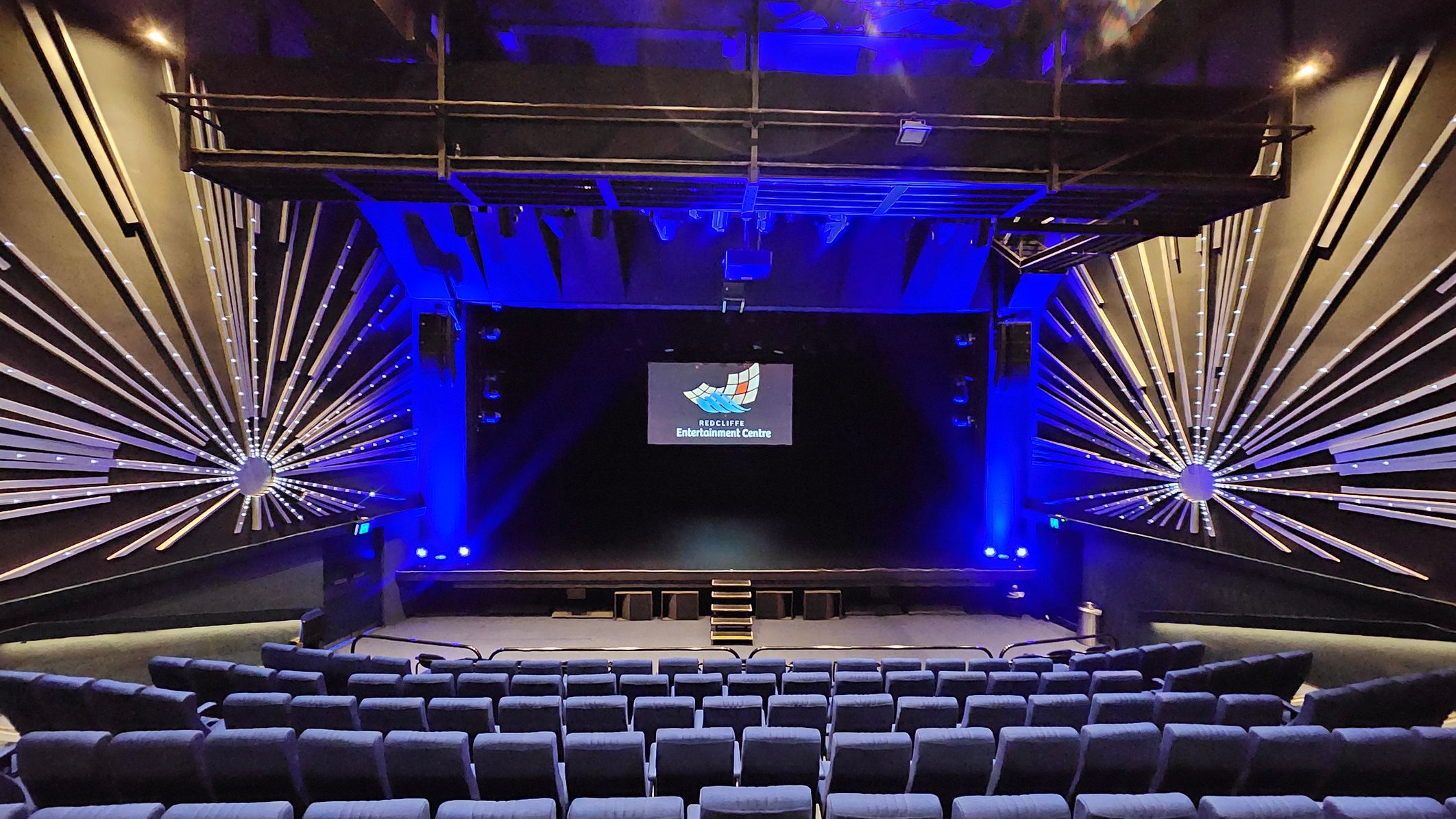 A theatre interior with rows of blue cushioned seats in the foreground. The stage is centred with a small set of steps leading up to it. Above the stage, a screen displays a logo and text. The walls on either side feature decorative radial patterns with illuminated lines extending outward. The ceiling is dark with blue lighting and visible structural elements. A theatre interior with rows of blue cushioned seats in the foreground. The stage is centred with a small set of steps leading up to it. Above the stage, a screen displays a logo and text. The walls on either side feature decorative radial patterns with illuminated lines extending outward. The ceiling is dark with blue lighting and visible structural elements.