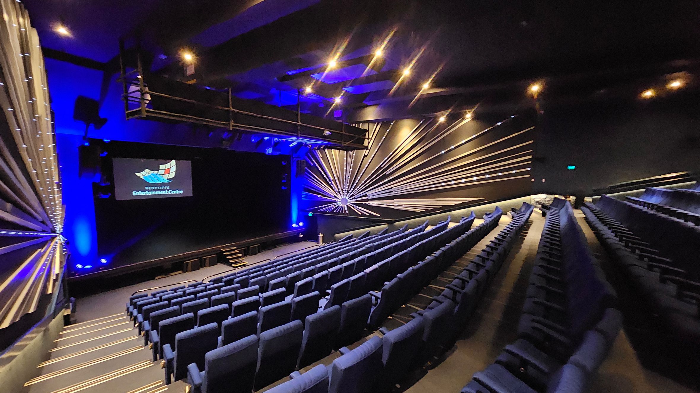 A theatre interior with rows of blue seats arranged in a tiered fashion on the right. The stage is at the front with a screen displaying "Redcliffe Entertainment Centre". The walls feature a decorative design with radiating lines and blue lighting. The ceiling has recessed lighting, and there is a balcony or technical area above the stage. The floor is carpeted with illuminated steps leading down to the stage. A theatre interior with rows of blue seats arranged in a tiered fashion on the right. The stage is at the front with a screen displaying "Redcliffe Entertainment Centre". The walls feature a decorative design with radiating lines and blue lighting. The ceiling has recessed lighting, and there is a balcony or technical area above the stage. The floor is carpeted with illuminated steps leading down to the stage.