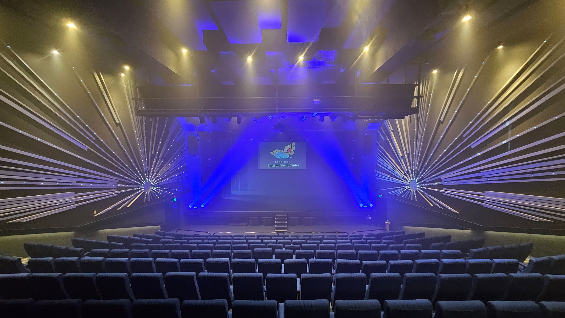 A theatre interior with rows of blue upholstered seats in the foreground. The stage is in the centre, illuminated by blue lights and featuring a screen displaying a logo. The walls on either side have decorative patterns with radiating lines, creating a starburst effect. The ceiling is equipped with spotlights, casting a warm glow. The overall atmosphere is enhanced by a misty effect, likely from stage smoke. A theatre interior with rows of blue upholstered seats in the foreground. The stage is in the centre, illuminated by blue lights and featuring a screen displaying a logo. The walls on either side have decorative patterns with radiating lines, creating a starburst effect. The ceiling is equipped with spotlights, casting a warm glow. The overall atmosphere is enhanced by a misty effect, likely from stage smoke.