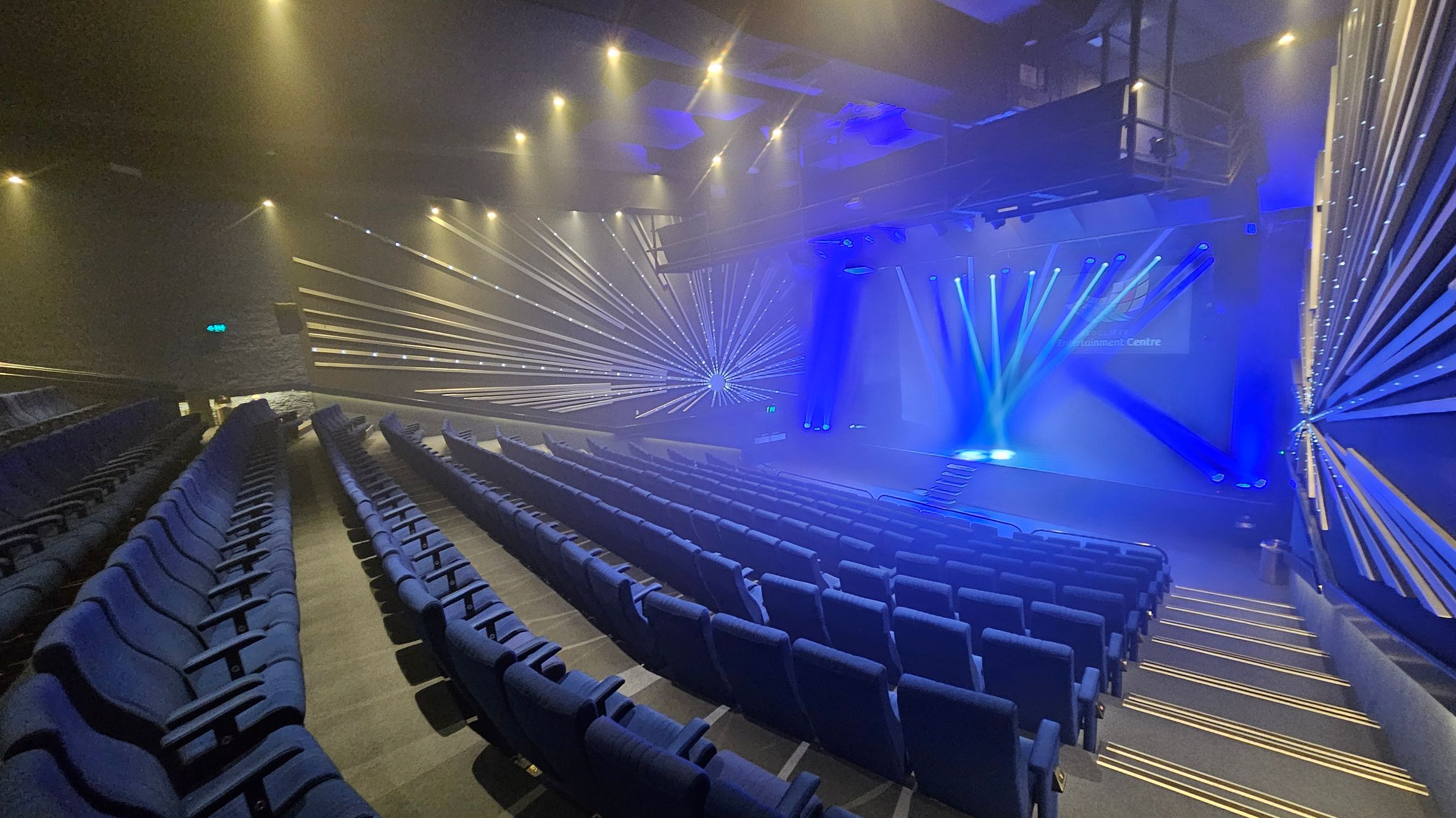 A theatre interior with rows of blue seats arranged in a semi-circular pattern. The stage is illuminated with blue spotlights, and beams of light radiate from the stage towards the audience. The walls have a geometric design with lines converging towards a central point. The ceiling is equipped with multiple spotlights, and the floor is carpeted with steps leading down to the stage. A theatre interior with rows of blue seats arranged in a semi-circular pattern. The stage is illuminated with blue spotlights, and beams of light radiate from the stage towards the audience. The walls have a geometric design with lines converging towards a central point. The ceiling is equipped with multiple spotlights, and the floor is carpeted with steps leading down to the stage.