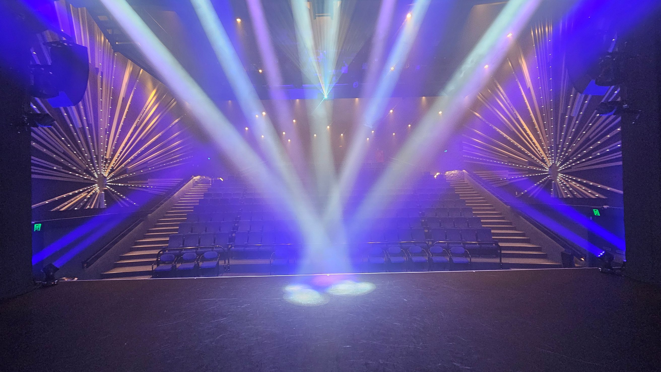 A theatre stage with bright, colourful spotlights shining towards the audience. The seating area is visible, with rows of empty seats. On both sides, there are decorative starburst patterns with illuminated lines extending outward. The ceiling is dark, with additional lights creating a vibrant atmosphere. The floor of the stage is dark and smooth. A theatre stage with bright, colourful spotlights shining towards the audience. The seating area is visible, with rows of empty seats. On both sides, there are decorative starburst patterns with illuminated lines extending outward. The ceiling is dark, with additional lights creating a vibrant atmosphere. The floor of the stage is dark and smooth.