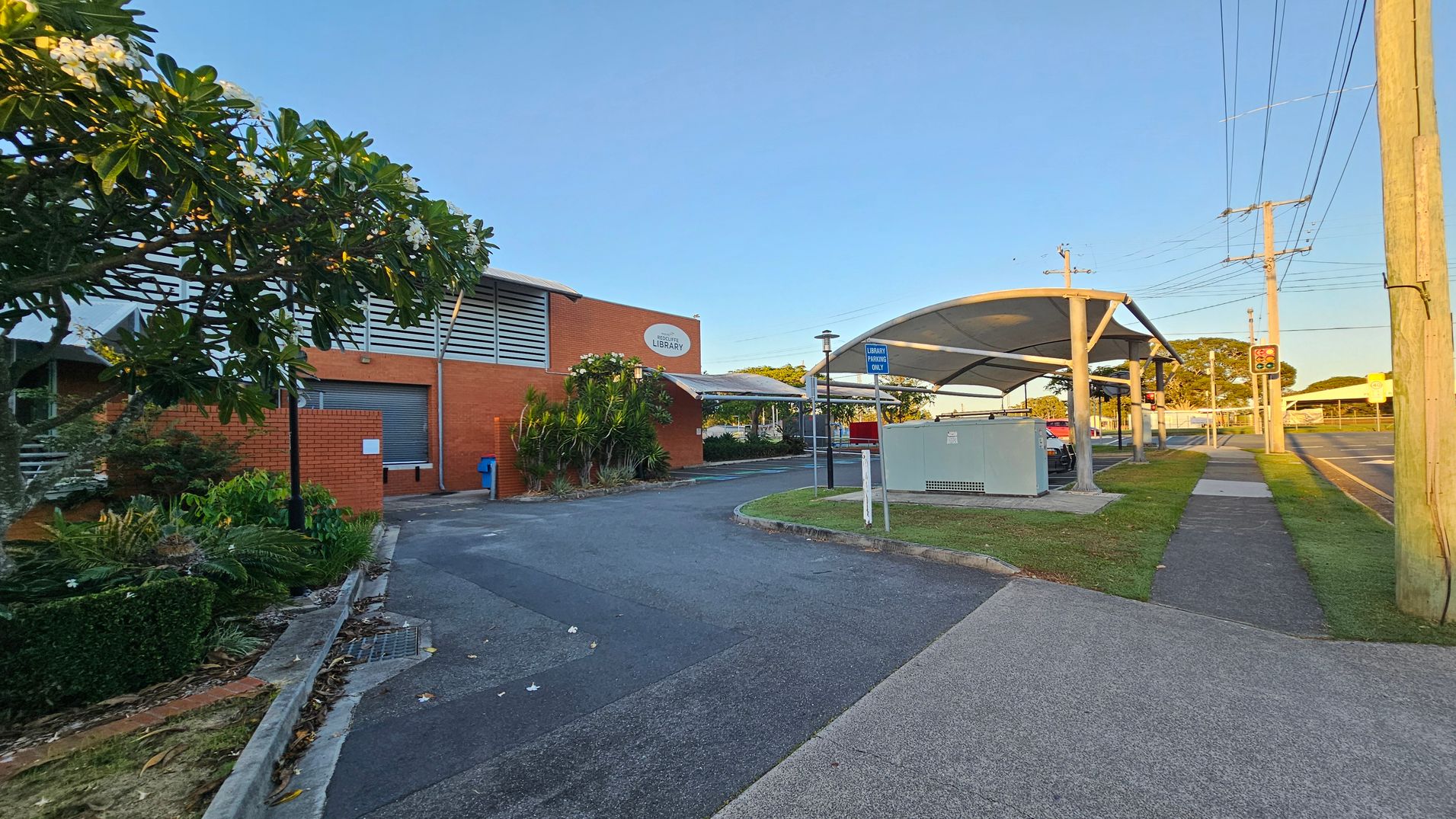 A brick building with a sign indicating it's a library is on the left, surrounded by lush greenery and a tree with white flowers. A paved driveway leads to the building. On the right, there's a covered walkway with a sign for library parking, bordered by a grassy area and a footpath. Utility poles and traffic lights are visible along the road in the background. The scene is well-lit, suggesting it's either early morning or late afternoon. A brick building with a sign indicating it's a library is on the left, surrounded by lush greenery and a tree with white flowers. A paved driveway leads to the building. On the right, there's a covered walkway with a sign for library parking, bordered by a grassy area and a footpath. Utility poles and traffic lights are visible along the road in the background. The scene is well-lit, suggesting it's either early morning or late afternoon.