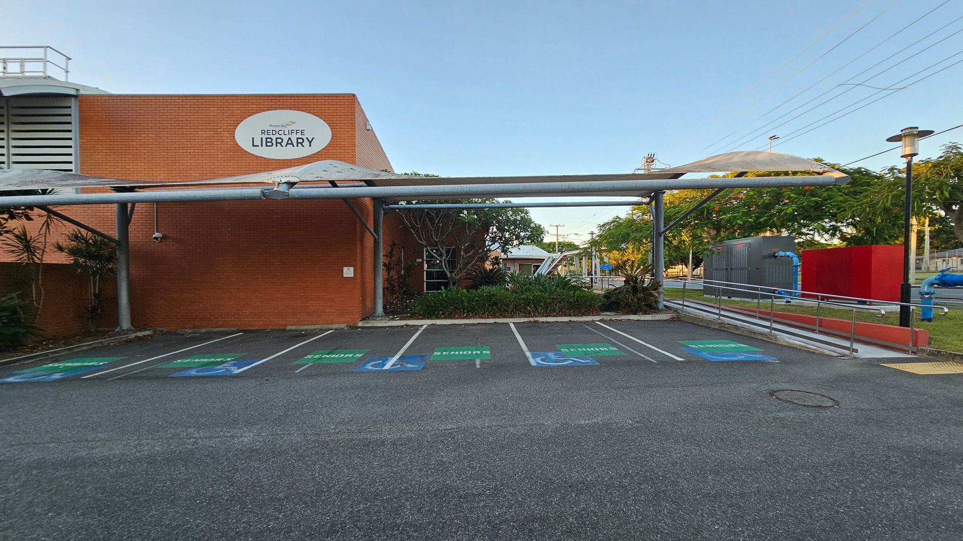 A brick building with a sign reading "Redcliffe Library" on the left. In front, there are several parking spaces marked for seniors and disabled access. A metal canopy extends over the parking area. On the right, there is a ramp with railings leading to the entrance, surrounded by greenery and trees. A red wall and some blue pipes are visible in the background, along with a streetlamp. The ground is asphalt, and the area is well-maintained. A brick building with a sign reading "Redcliffe Library" on the left. In front, there are several parking spaces marked for seniors and disabled access. A metal canopy extends over the parking area. On the right, there is a ramp with railings leading to the entrance, surrounded by greenery and trees. A red wall and some blue pipes are visible in the background, along with a streetlamp. The ground is asphalt, and the area is well-maintained.