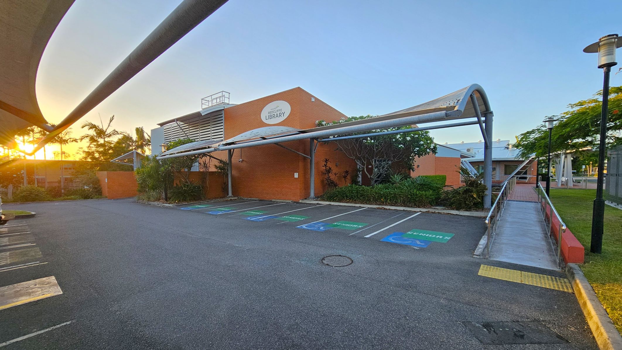 A parking area with designated spaces for seniors is in the foreground, marked with green and blue paint. A covered walkway leads to a brick building with a sign reading "Redcliffe Library" in the centre. The building is surrounded by greenery, including palm trees and shrubs. On the right, a ramp with metal railings leads to the entrance. The sun is setting on the left, casting a warm glow over the scene. A streetlamp is visible on the right, and the sky is clear. A parking area with designated spaces for seniors is in the foreground, marked with green and blue paint. A covered walkway leads to a brick building with a sign reading "Redcliffe Library" in the centre. The building is surrounded by greenery, including palm trees and shrubs. On the right, a ramp with metal railings leads to the entrance. The sun is setting on the left, casting a warm glow over the scene. A streetlamp is visible on the right, and the sky is clear.
