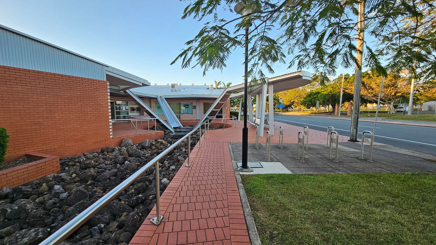 A brick pathway leads to a building with a red brick exterior on the left. The path is bordered by a metal railing and a bed of dark rocks. In the centre, a modern structure with angled metal beams and a glass entrance is visible. On the right, there are bike racks and a grassy area. A road runs parallel to the building, lined with trees and a utility pole. The sky is clear, and the area is well-lit. A brick pathway leads to a building with a red brick exterior on the left. The path is bordered by a metal railing and a bed of dark rocks. In the centre, a modern structure with angled metal beams and a glass entrance is visible. On the right, there are bike racks and a grassy area. A road runs parallel to the building, lined with trees and a utility pole. The sky is clear, and the area is well-lit.