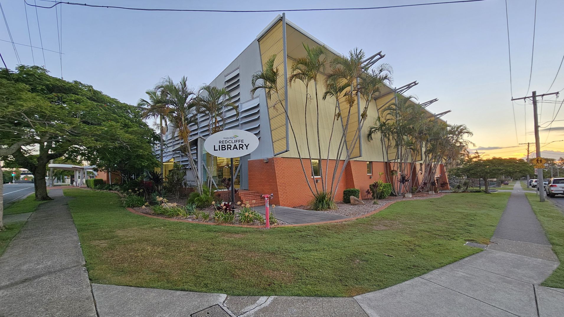 A modern building with a slanted roof and a mix of brick and metal siding, identified as "Redcliffe Library" on a sign in front. Tall palm trees line the side of the building. The foreground features a grassy area with a concrete footpath leading to the entrance. On the left, a large tree provides shade over the footpath, which runs parallel to a road. On the right, a row of parked cars is visible along the street. Power lines stretch across the top of the image. A modern building with a slanted roof and a mix of brick and metal siding, identified as "Redcliffe Library" on a sign in front. Tall palm trees line the side of the building. The foreground features a grassy area with a concrete footpath leading to the entrance. On the left, a large tree provides shade over the footpath, which runs parallel to a road. On the right, a row of parked cars is visible along the street. Power lines stretch across the top of the image.