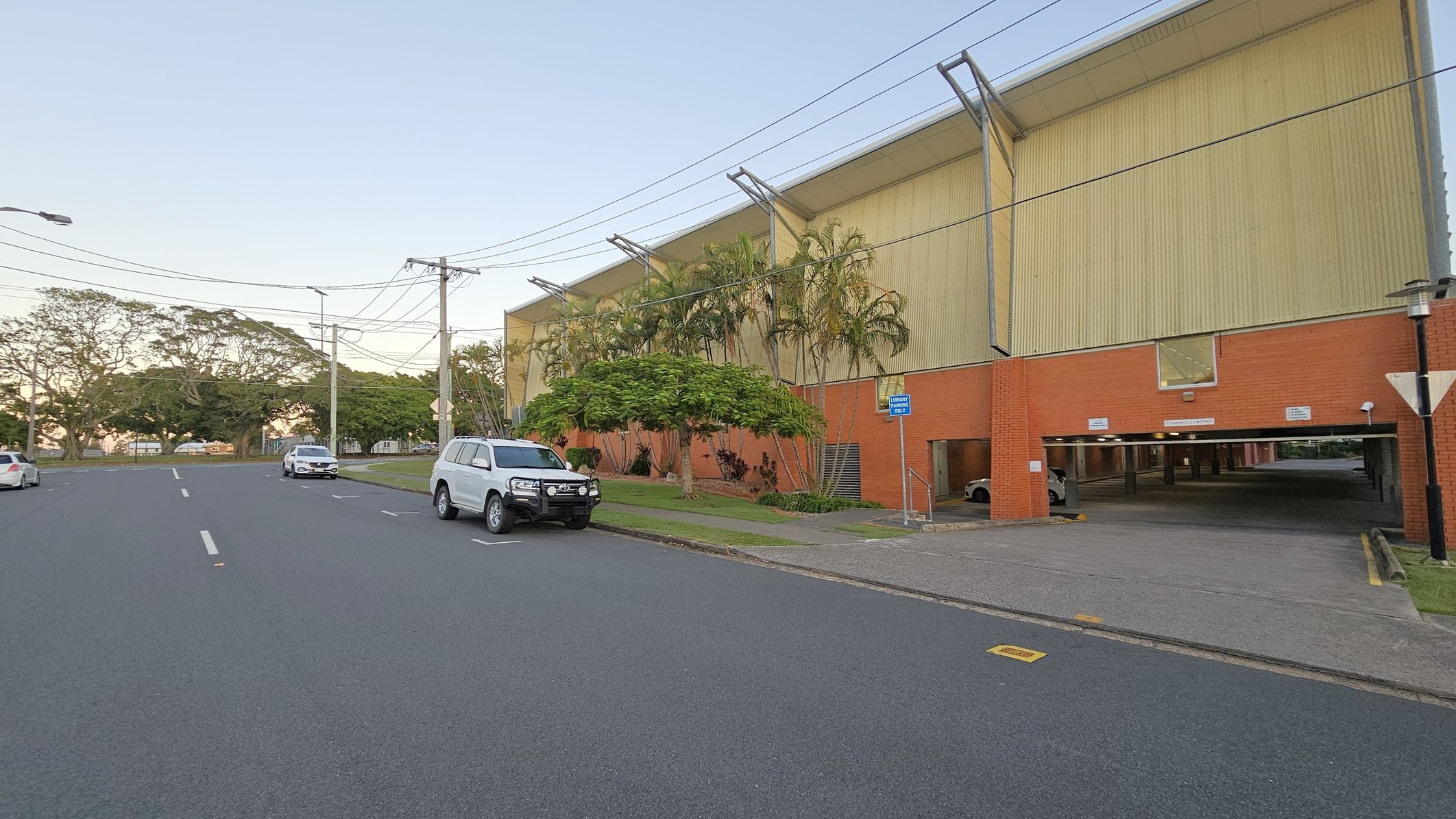 A street scene with a large building on the right, featuring a red brick lower section and a beige upper section. Several palm trees and shrubs are in front of the building. A white SUV is parked on the street, with other cars visible in the distance. The road is wide with marked lanes, and power lines run overhead. The background includes more trees and a clear sky. A covered parking area is visible under the building. A street scene with a large building on the right, featuring a red brick lower section and a beige upper section. Several palm trees and shrubs are in front of the building. A white SUV is parked on the street, with other cars visible in the distance. The road is wide with marked lanes, and power lines run overhead. The background includes more trees and a clear sky. A covered parking area is visible under the building.