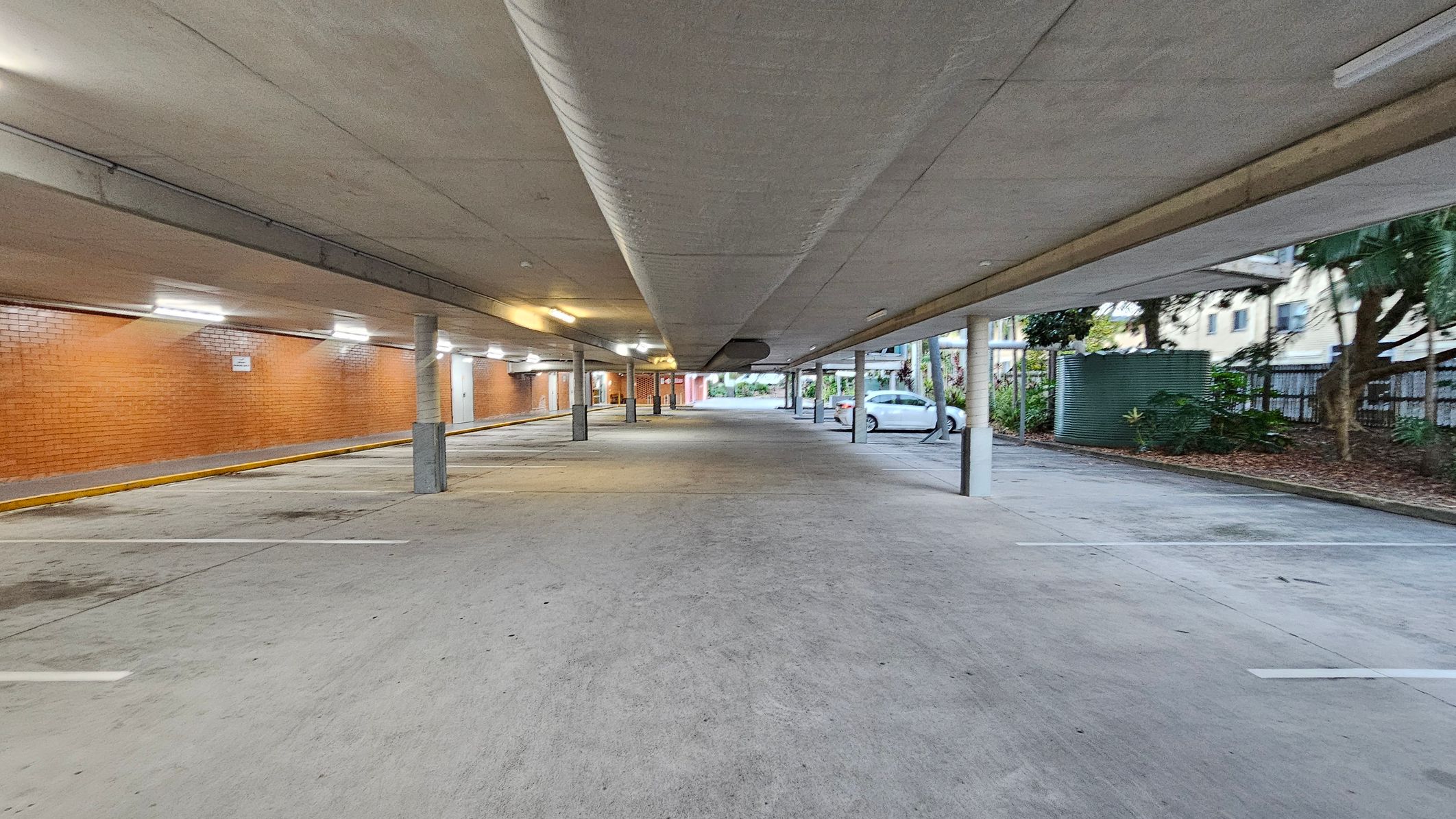 A covered parking area with a concrete floor and ceiling. On the left, there is a long brick wall with several lights illuminating the space. The right side features a row of parking spaces, with a white car parked near some greenery and a large cylindrical water tank. Several support columns run down the centre, and the area is open at the far end, leading to an outdoor space with trees and buildings visible in the background. A covered parking area with a concrete floor and ceiling. On the left, there is a long brick wall with several lights illuminating the space. The right side features a row of parking spaces, with a white car parked near some greenery and a large cylindrical water tank. Several support columns run down the centre, and the area is open at the far end, leading to an outdoor space with trees and buildings visible in the background.