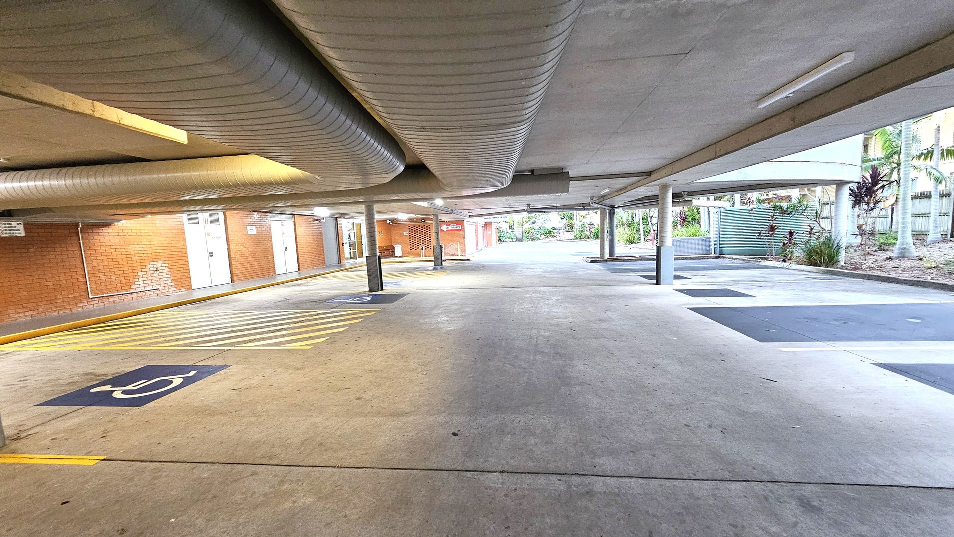 A covered parking area with a concrete floor and ceiling. On the left, there are brick walls with several doors and a sign. Yellow lines and a disabled parking symbol are painted on the ground. Large ventilation ducts run along the ceiling. On the right, there are open spaces with some greenery and palm trees visible outside. Several support columns are evenly spaced throughout the area. A covered parking area with a concrete floor and ceiling. On the left, there are brick walls with several doors and a sign. Yellow lines and a disabled parking symbol are painted on the ground. Large ventilation ducts run along the ceiling. On the right, there are open spaces with some greenery and palm trees visible outside. Several support columns are evenly spaced throughout the area.