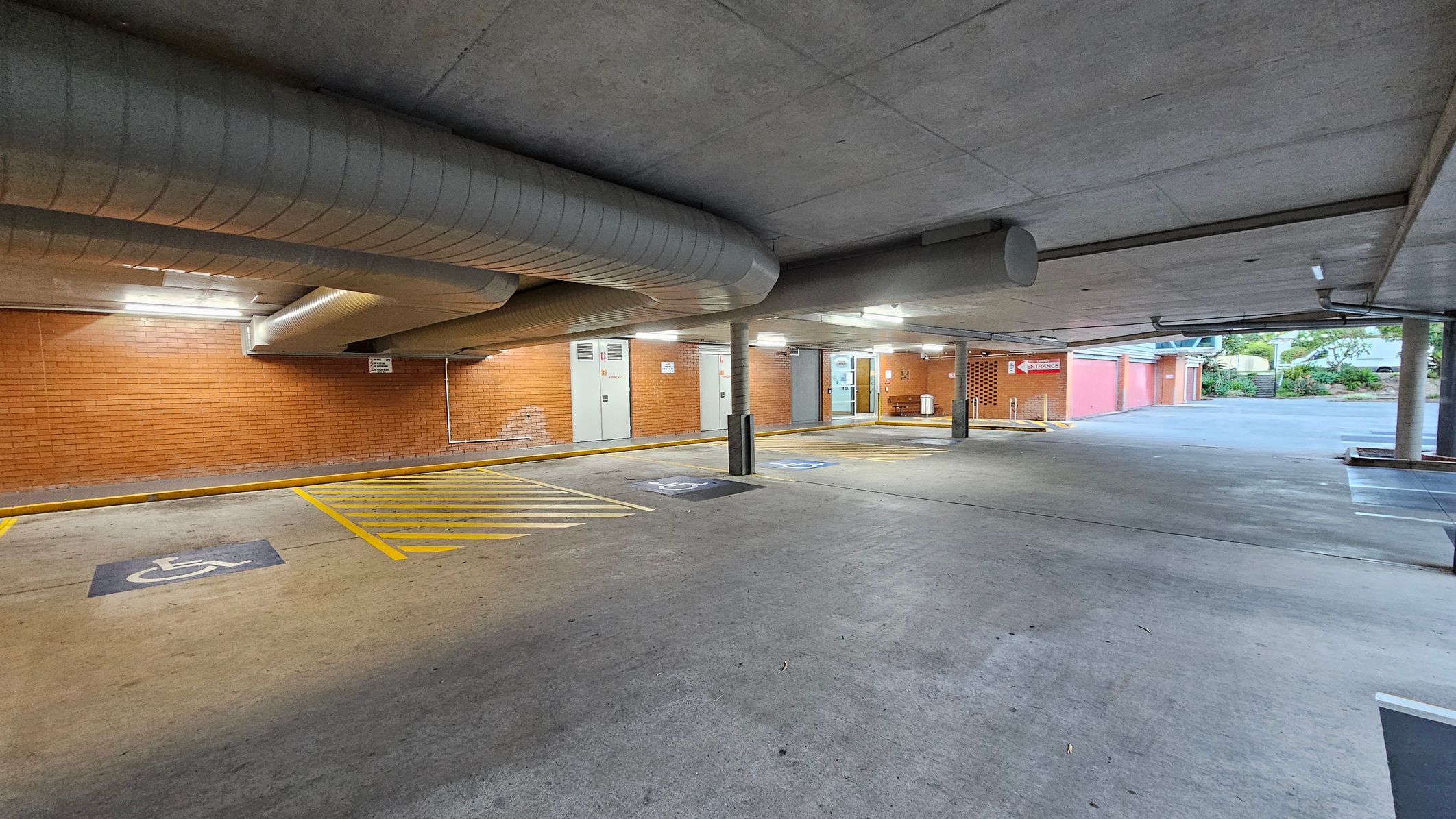 Underground car park with a concrete floor and ceiling. On the left, there are designated disabled parking spaces marked with yellow lines and symbols. The brick wall in the background has several doors and a sign indicating an entrance. Large ventilation ducts run along the ceiling. On the right, the car park opens to an outdoor area with greenery and a staircase leading upwards. The lighting is provided by overhead fluorescent lights. Underground car park with a concrete floor and ceiling. On the left, there are designated disabled parking spaces marked with yellow lines and symbols. The brick wall in the background has several doors and a sign indicating an entrance. Large ventilation ducts run along the ceiling. On the right, the car park opens to an outdoor area with greenery and a staircase leading upwards. The lighting is provided by overhead fluorescent lights.