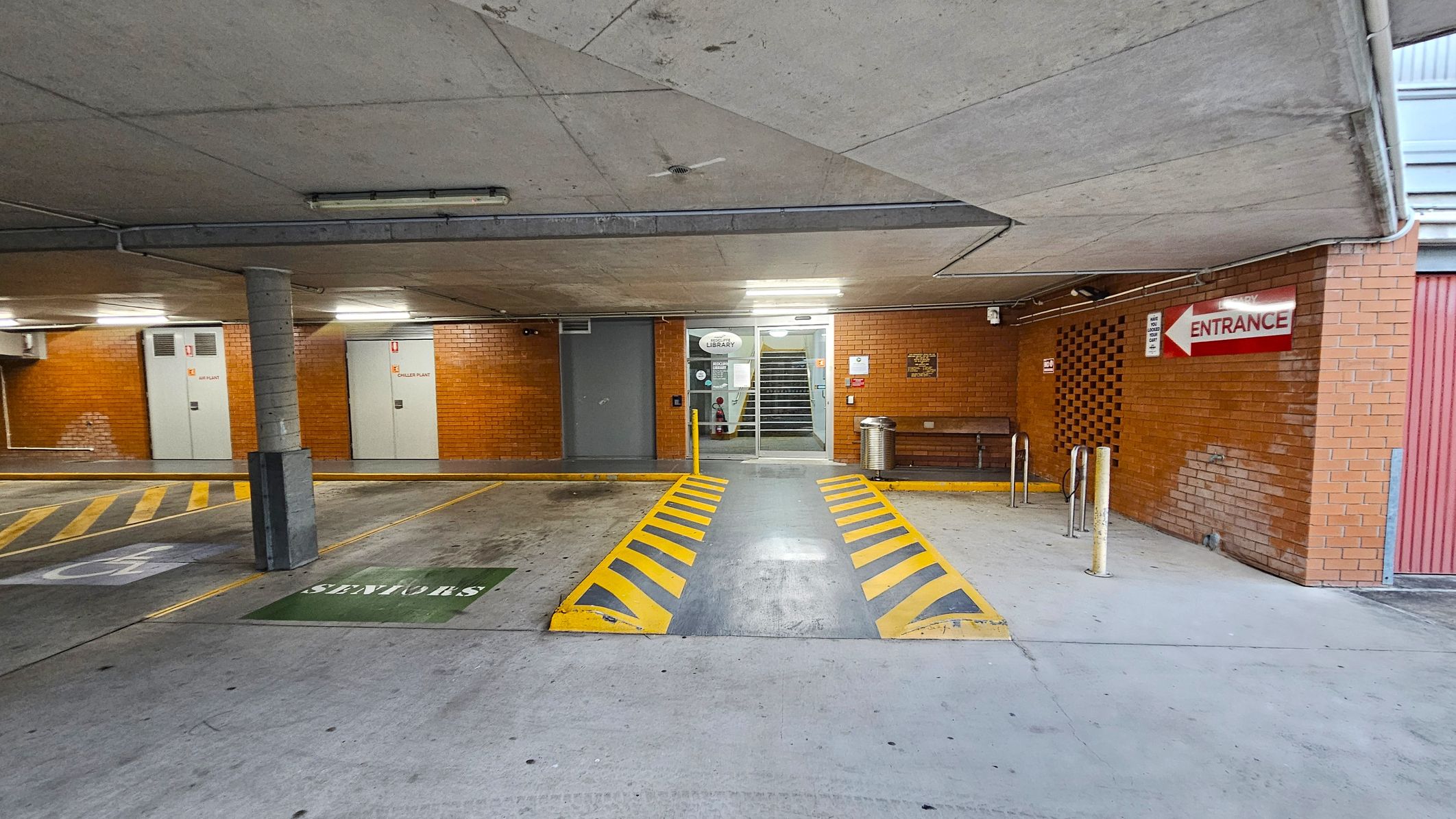 A concrete parking area with a ramp in the centre marked by yellow and black stripes. On the left, there are parking spaces with a "Seniors" sign and a pillar. The background features brick walls with doors labelled "Diesel Plant" and "Library" near a staircase. On the right, a red sign indicates "Entrance" above a bench and a metal bin. The ceiling is concrete with fluorescent lights. A concrete parking area with a ramp in the centre marked by yellow and black stripes. On the left, there are parking spaces with a "Seniors" sign and a pillar. The background features brick walls with doors labelled "Diesel Plant" and "Library" near a staircase. On the right, a red sign indicates "Entrance" above a bench and a metal bin. The ceiling is concrete with fluorescent lights.