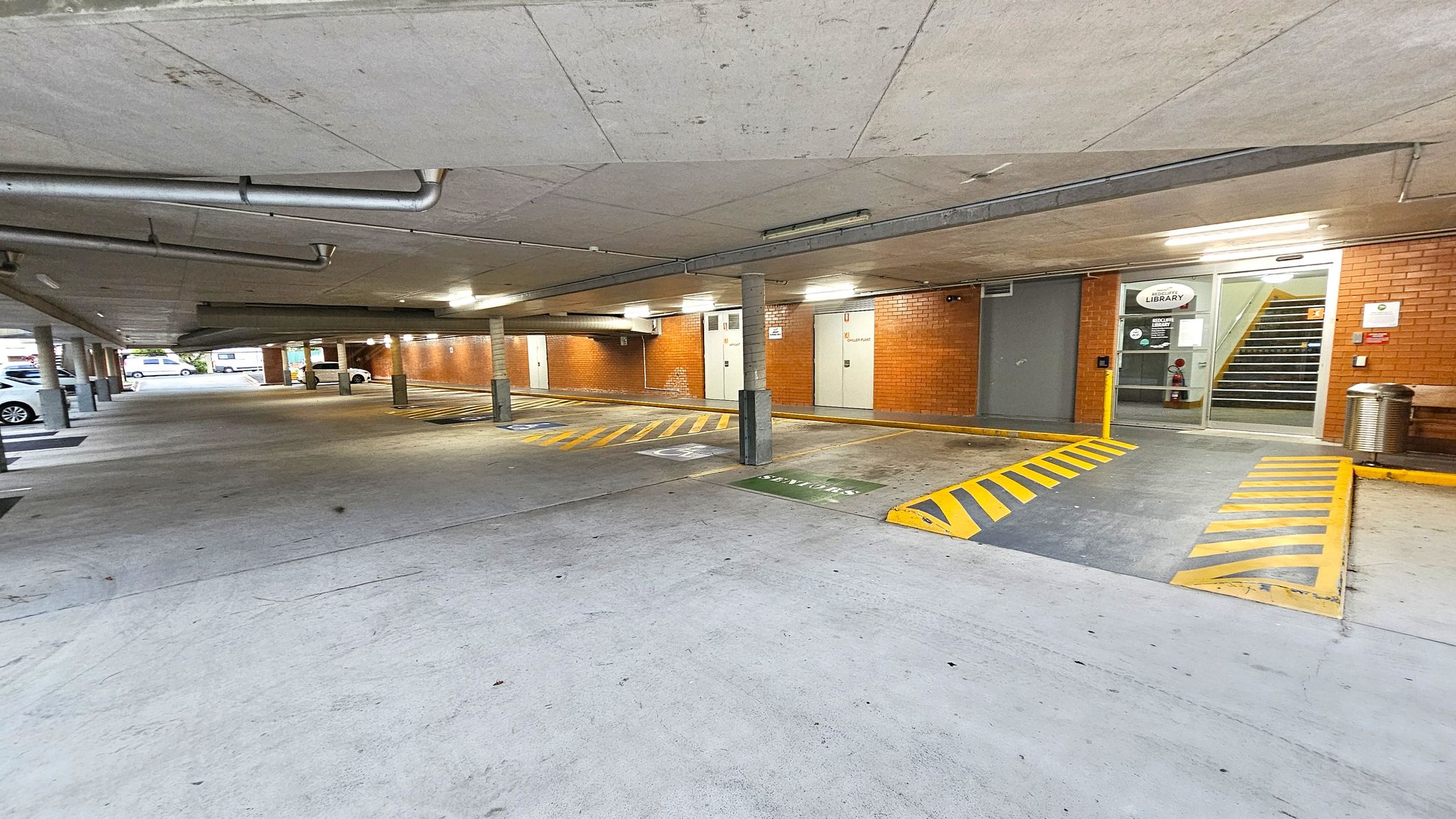 A covered car park with a concrete floor and ceiling. On the right, there is a brick wall with a glass door leading to a staircase, marked with a "Library" sign. Yellow and black striped markings are on the ground near the entrance. Several parking spaces are visible, with a few cars parked on the left. The area is lit with fluorescent lights, and there are metal pipes running along the ceiling. A metal bin and a bench are near the entrance. A covered car park with a concrete floor and ceiling. On the right, there is a brick wall with a glass door leading to a staircase, marked with a "Library" sign. Yellow and black striped markings are on the ground near the entrance. Several parking spaces are visible, with a few cars parked on the left. The area is lit with fluorescent lights, and there are metal pipes running along the ceiling. A metal bin and a bench are near the entrance.